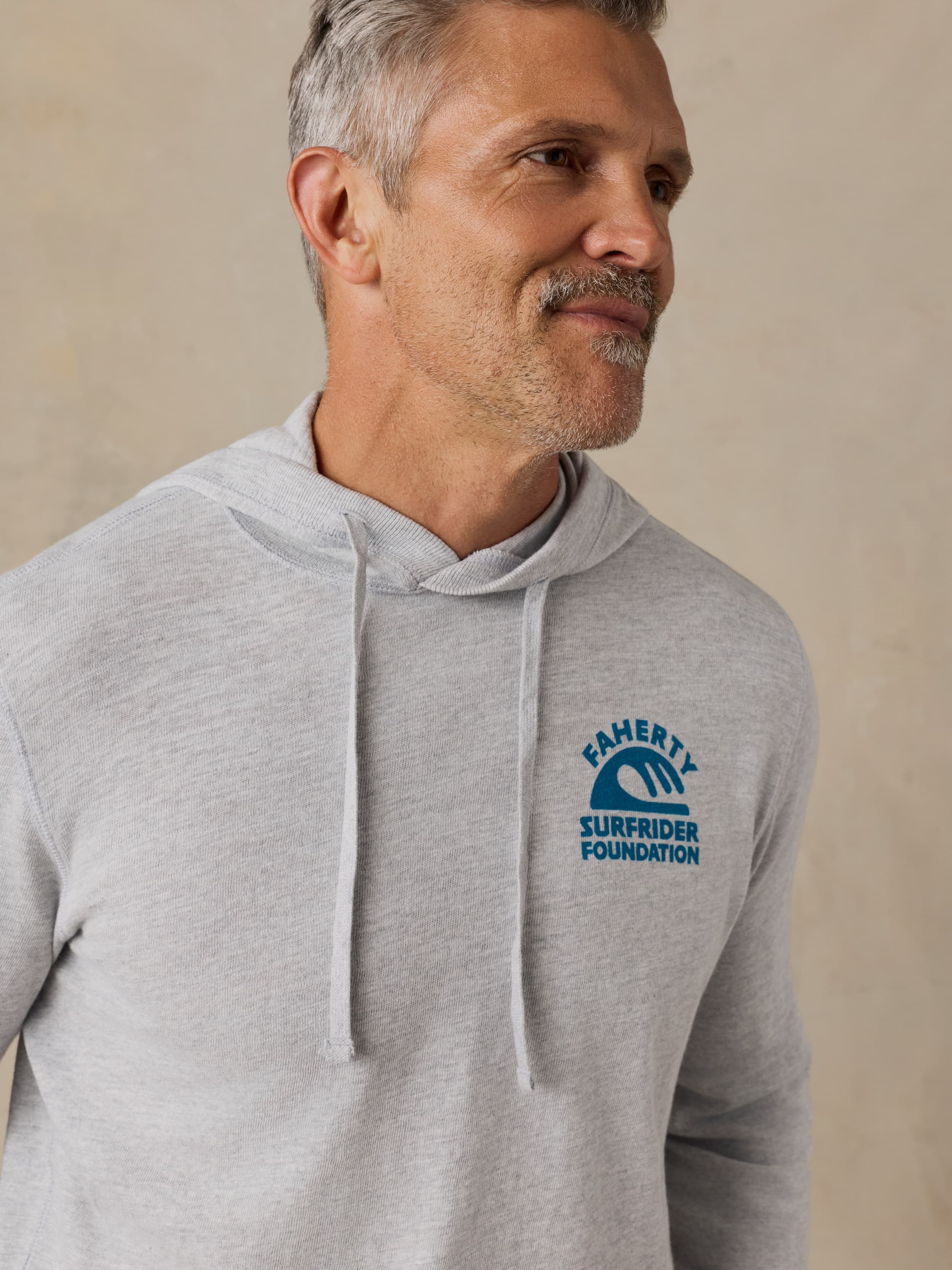 Man wearing a gray hoodie with a Faherty x Surfrider Foundation logo on a beige background