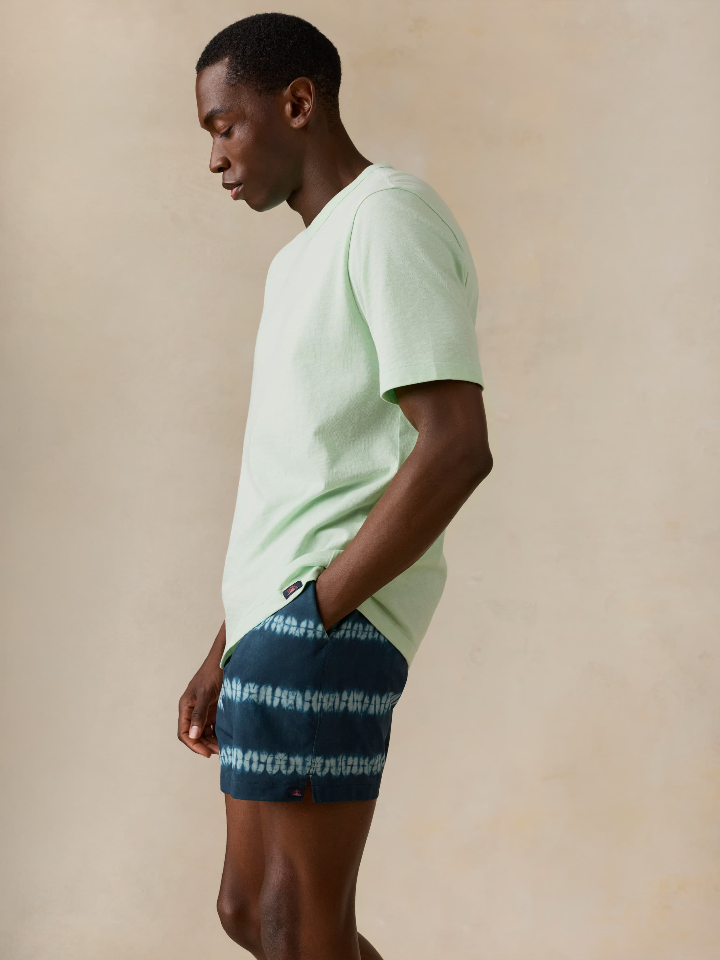 Man wearing a light green t-shirt and dark blue tie dye swim trunks on a beige background