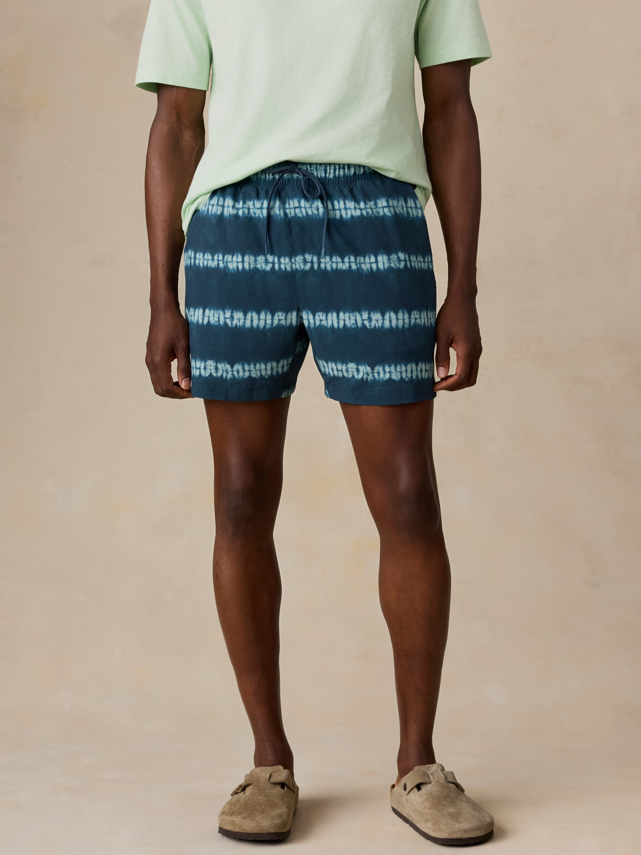 Man wearing blue tie-dye swim trunks and a light green shirt on a beige background