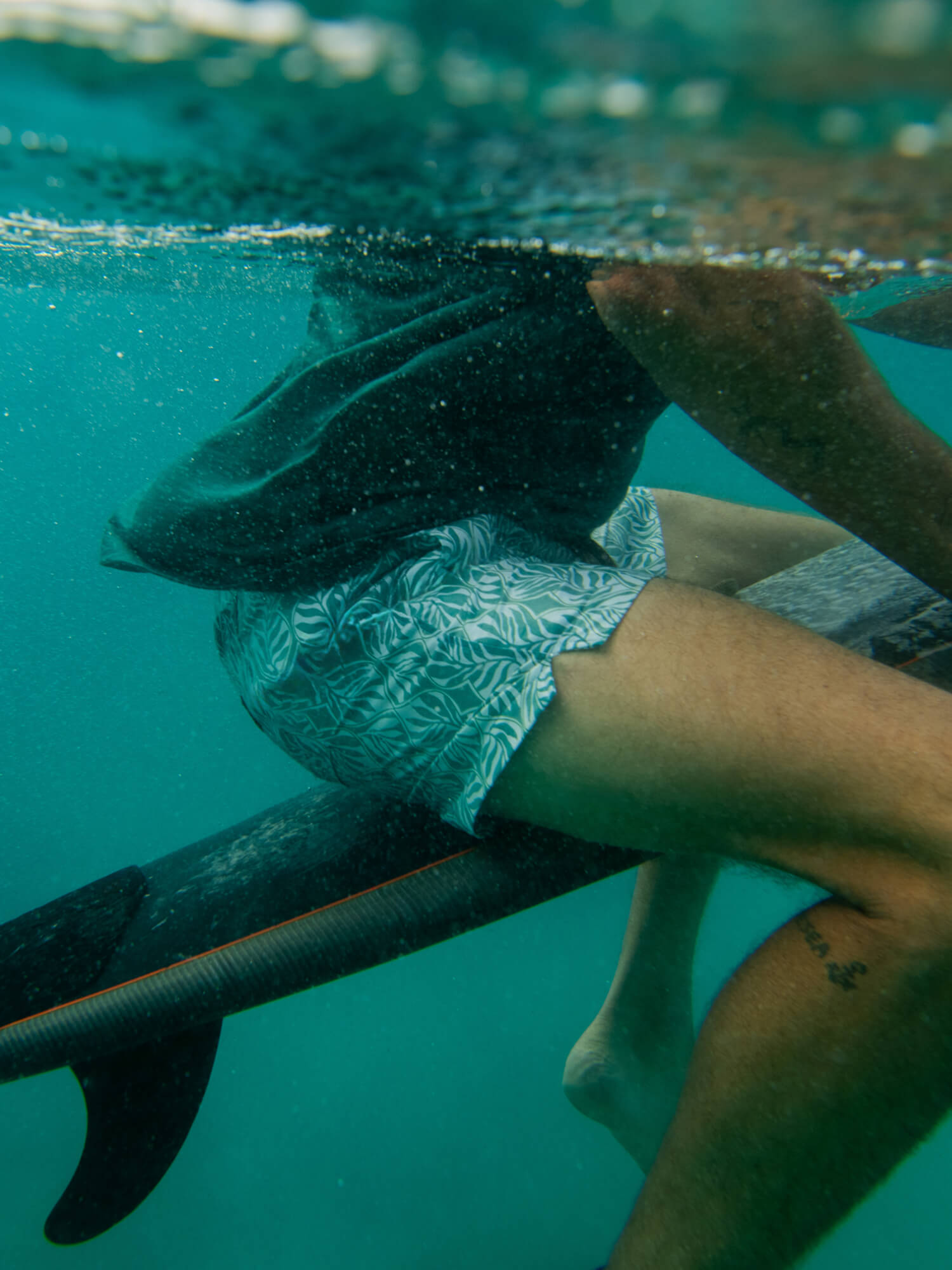 Man underwater with fins and a surfboard, wearing patterned shorts.