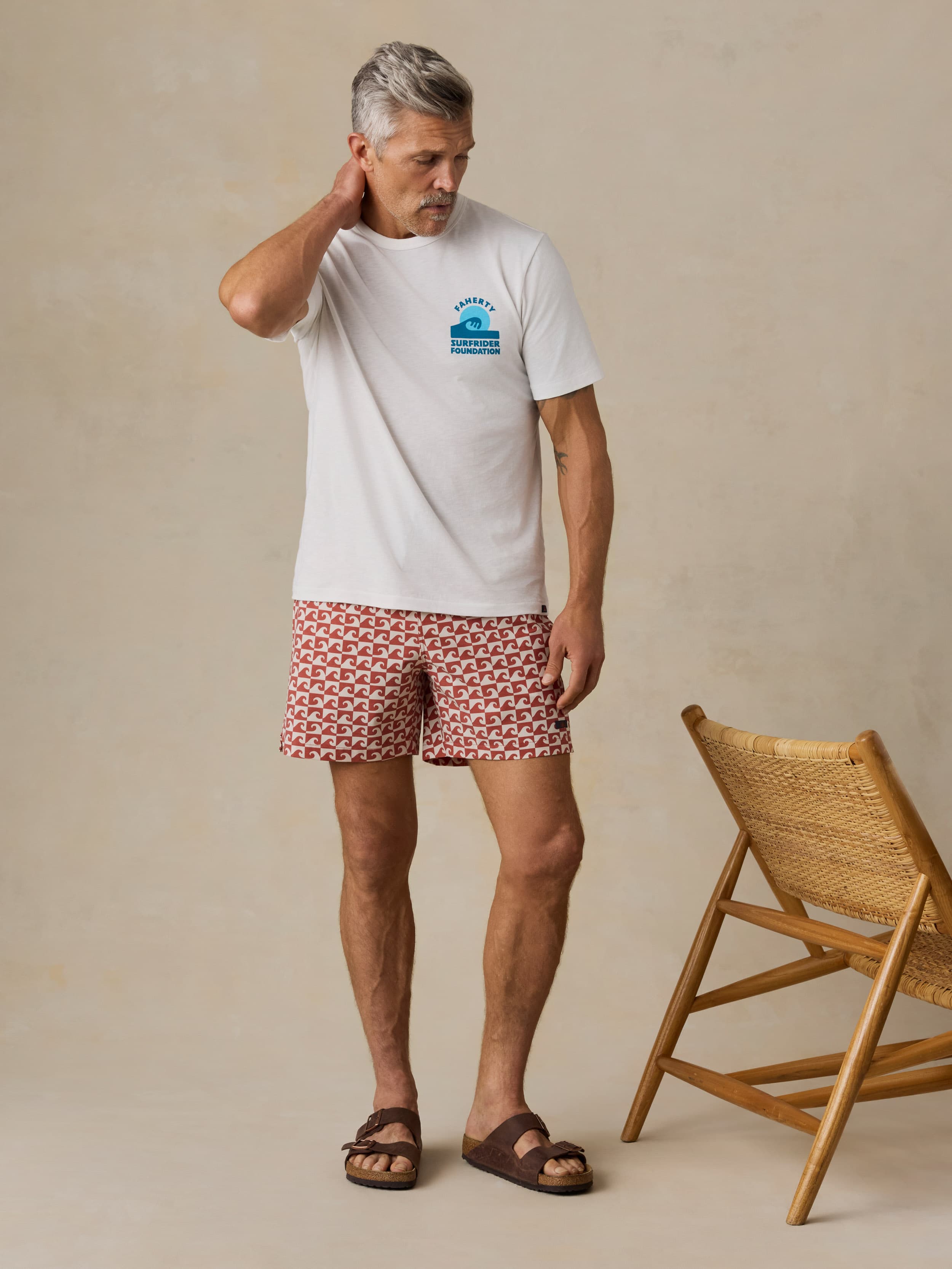 Man wearing a white t-shirt with a logo and red patterned swim shorts standing next to a wooden chair.