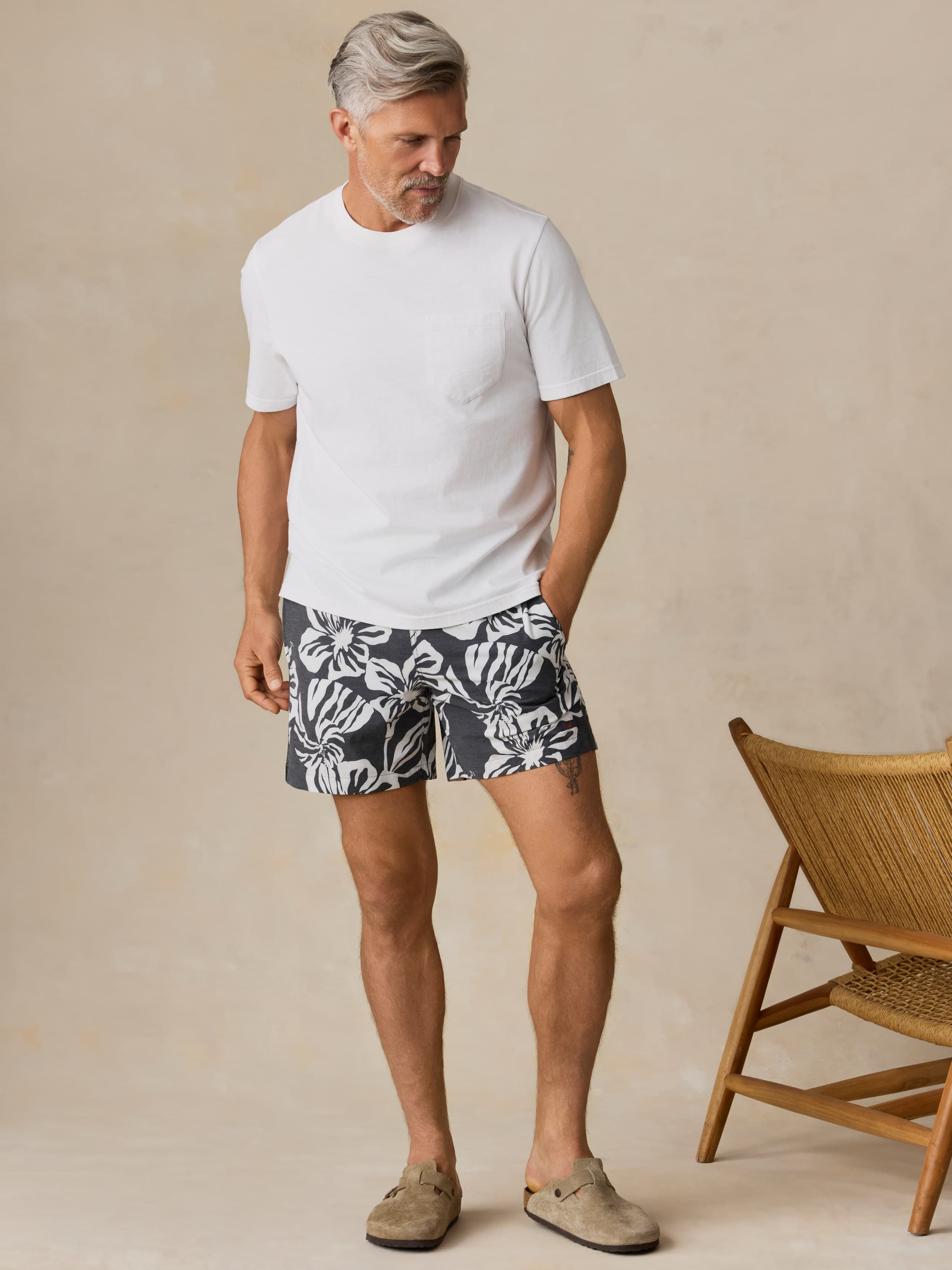 Man wearing a white t-shirt and floral patterned swim shorts standing against a beige wall with a wooden chair.