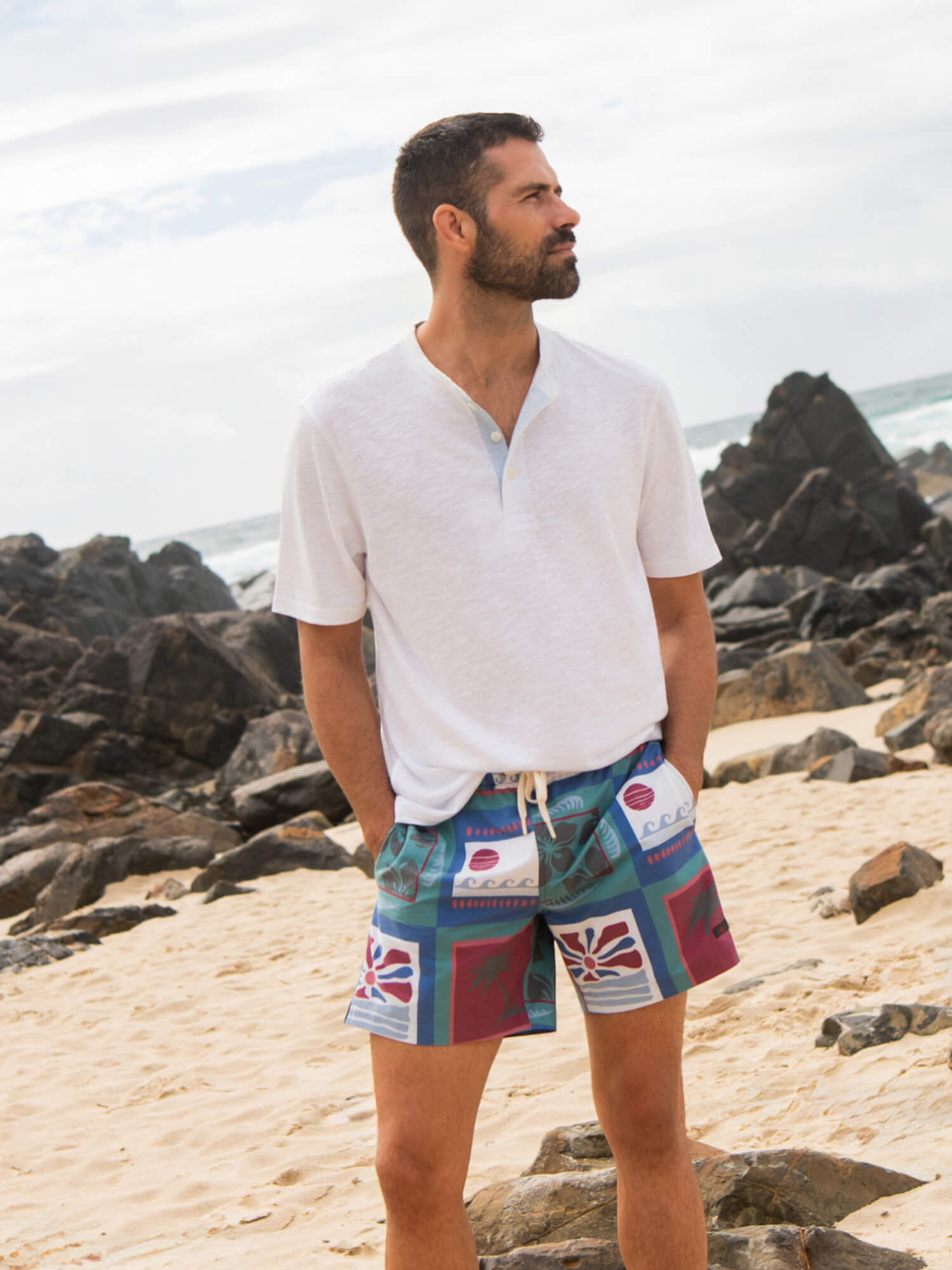 Man wearing a white henley and printed swim trunks at the beach