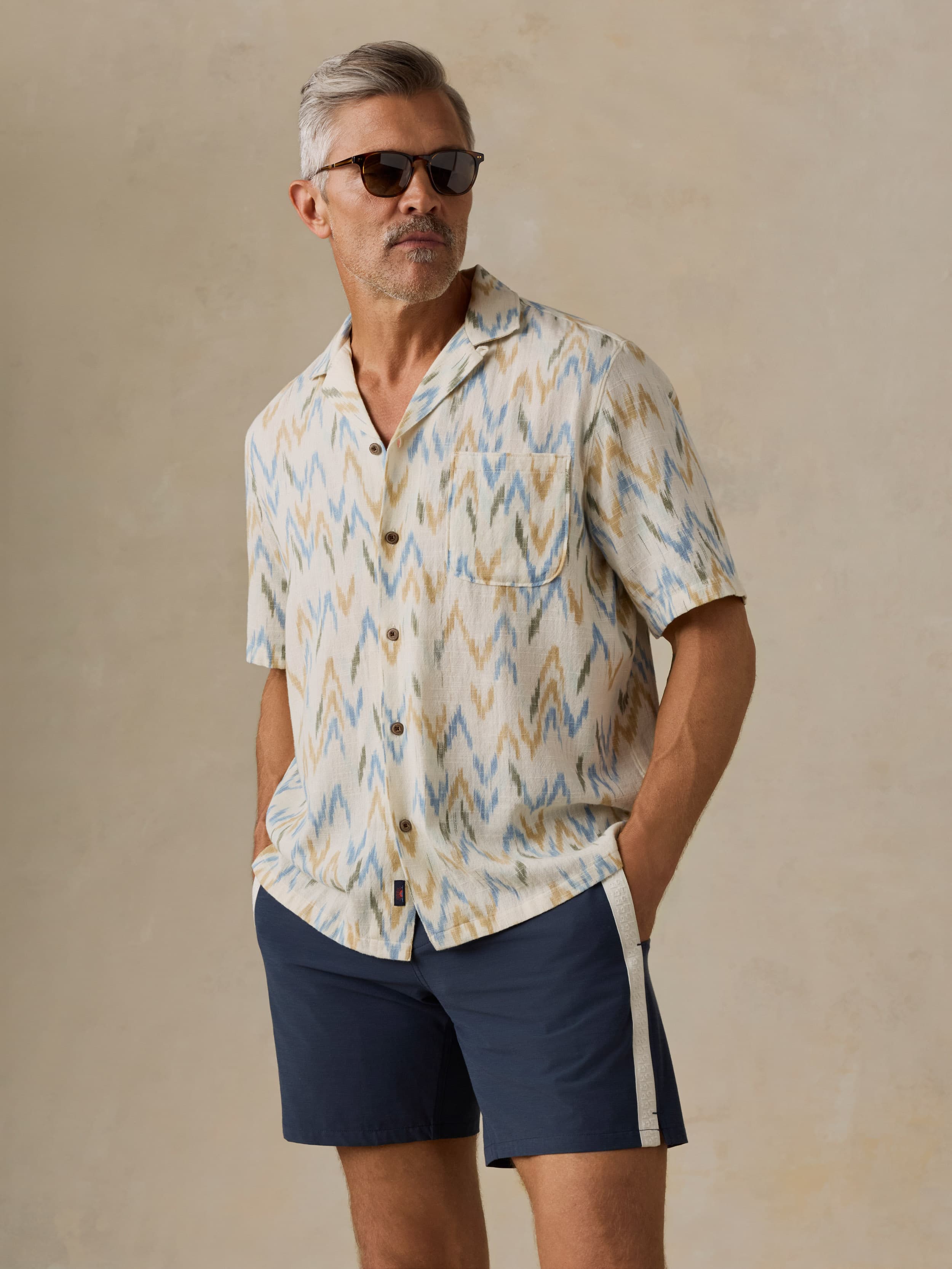 Man wearing a patterned shirt and navy swim shorts against a beige background
