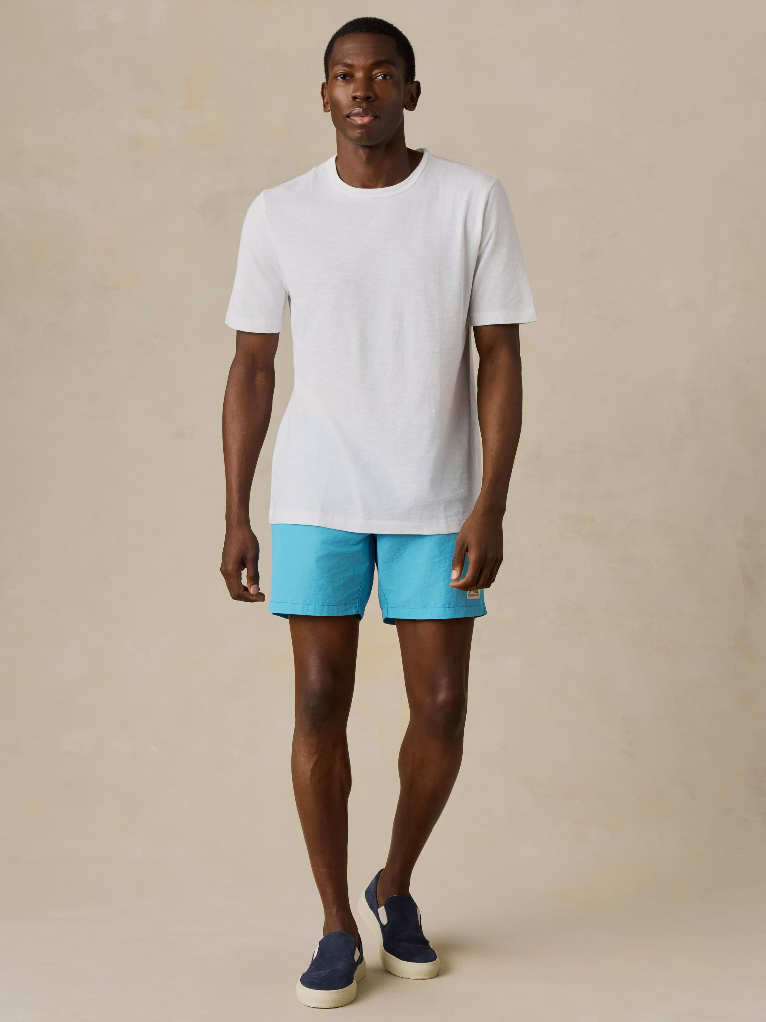 Man wearing a white t-shirt and blue swim shorts against a beige background