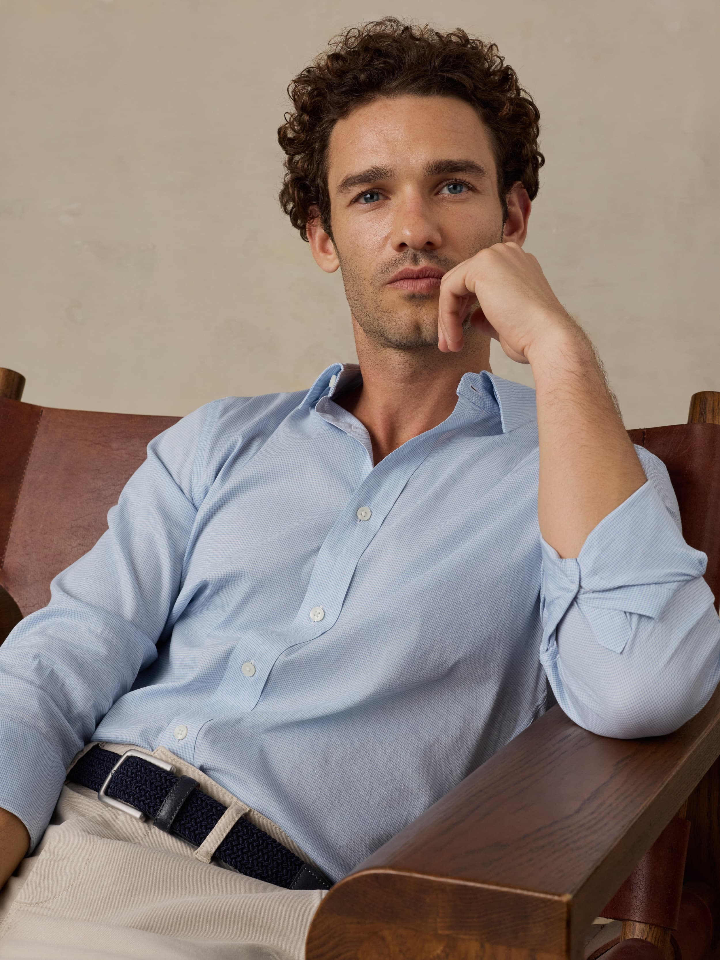 Man wearing a light blue shirt sitting on a wooden chair against a beige background