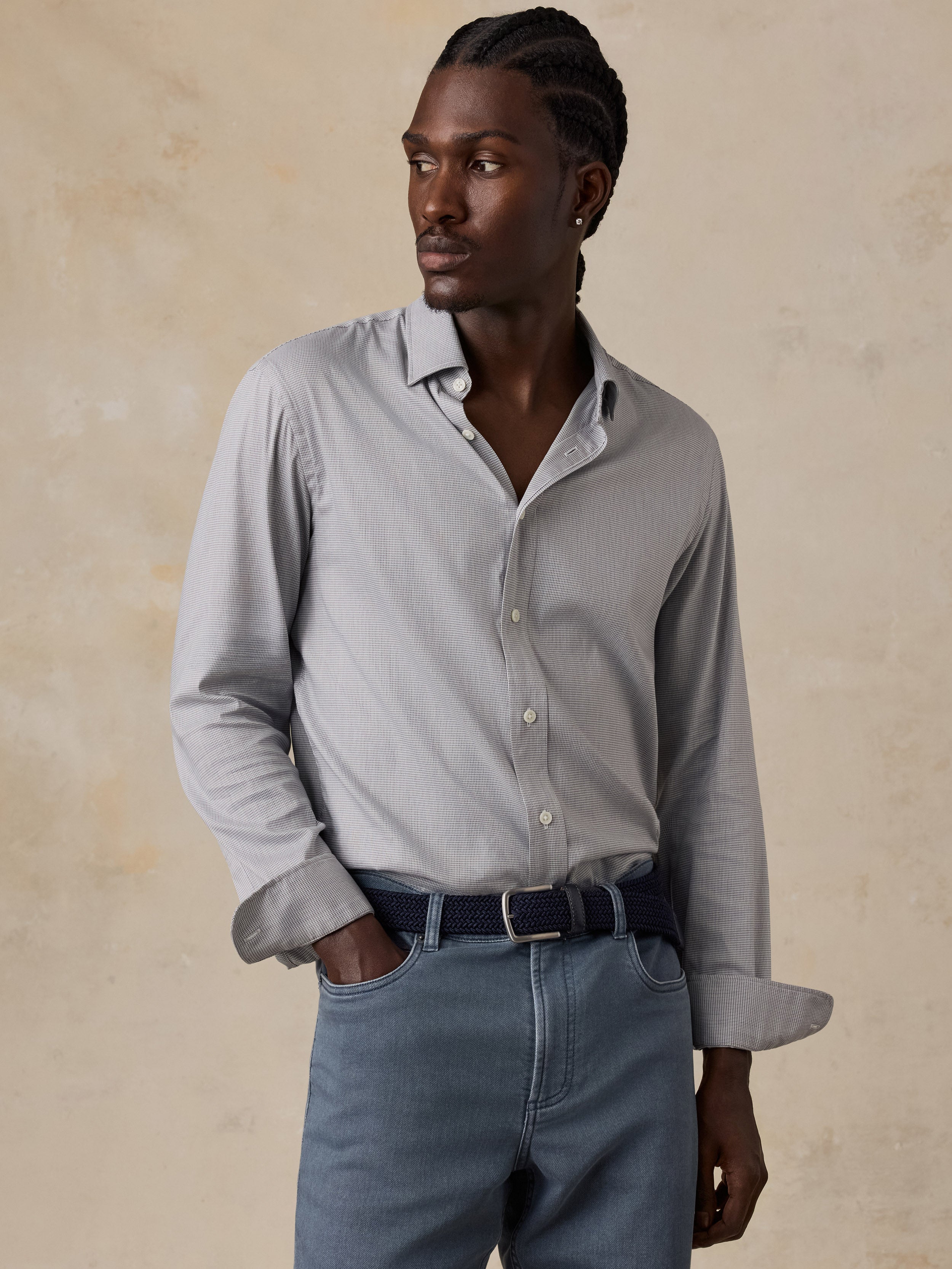 A man wearing a light gray and white vertically striped long-sleeve button-down shirt, paired with blue jeans and a dark belt, standing against a textured beige background.