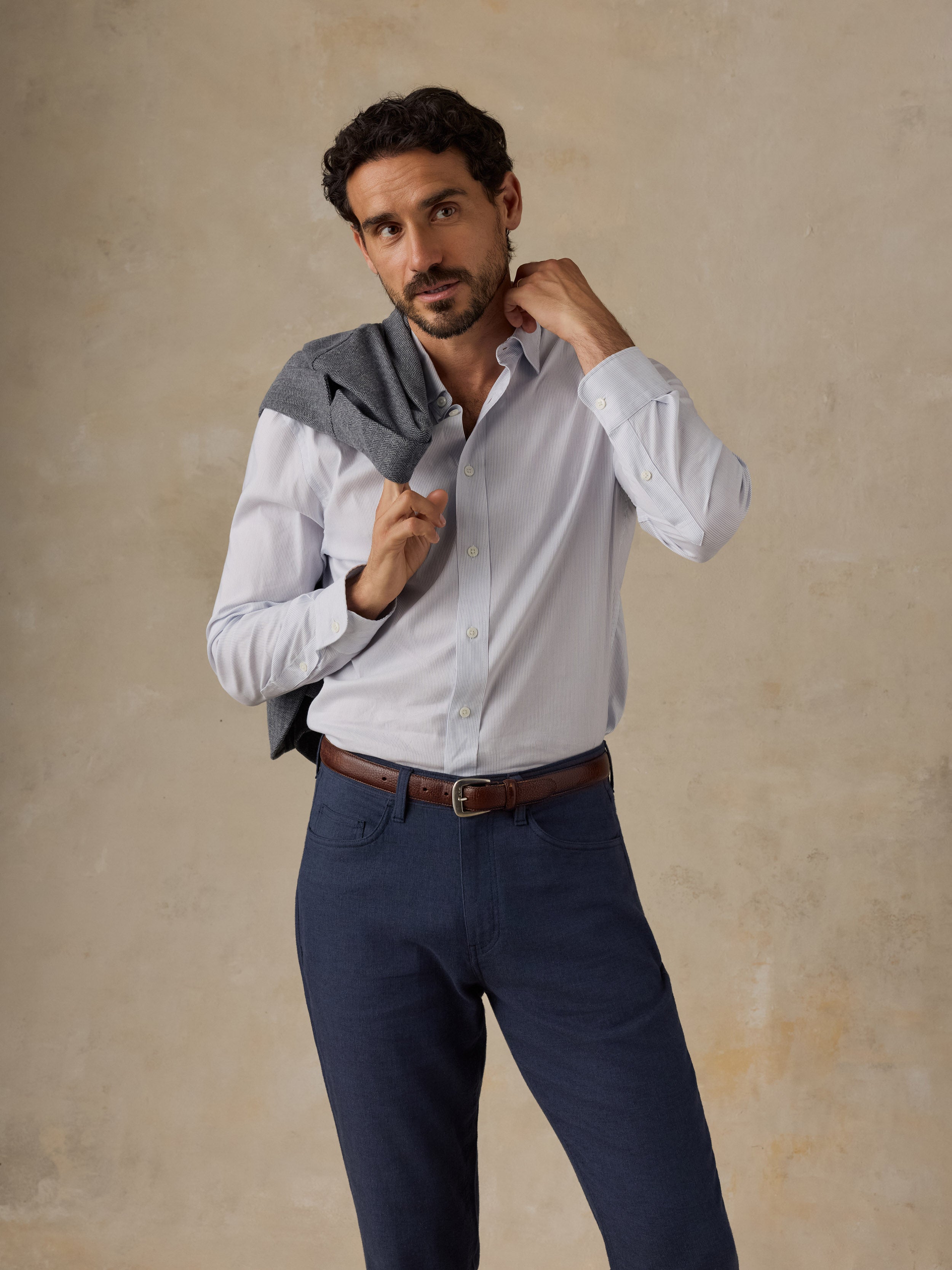 A man wearing a white long-sleeve button-down shirt and navy blue trousers, with a gray blazer draped over his shoulders, posing against a textured beige studio backdrop.