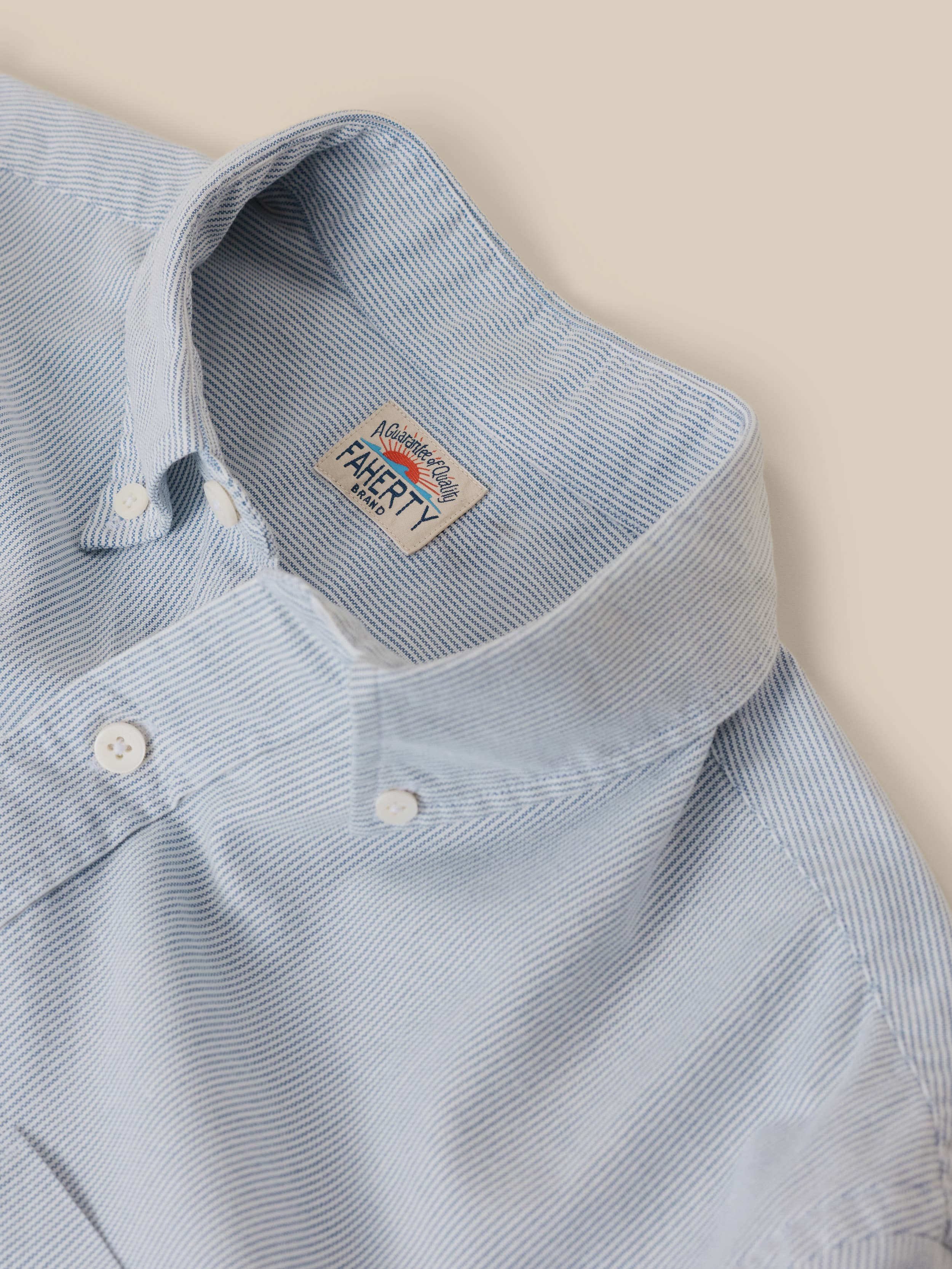 Close-up of a button down shirt with blue and white stripes and a Faherty brand logo at the neck against a beige background