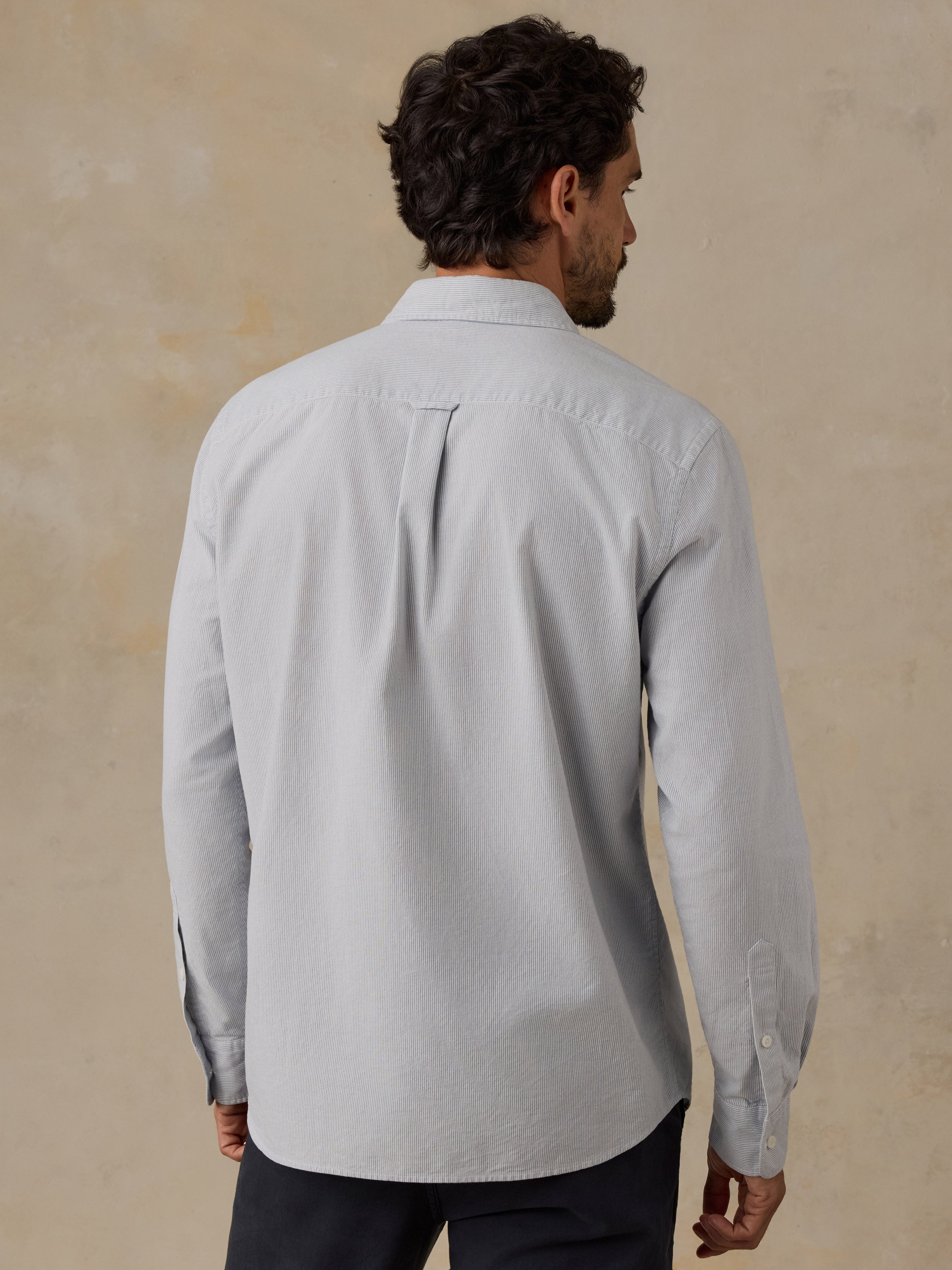 A man viewed from the back wearing a light blue long-sleeve button-down shirt and dark trousers, standing against a textured beige wall.