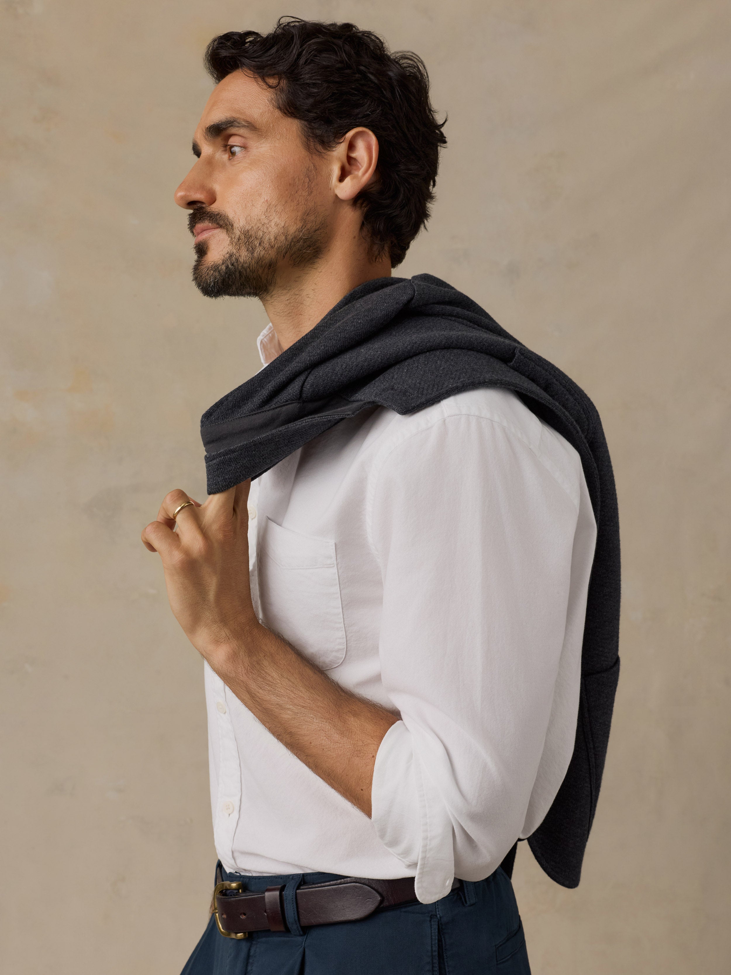 A man in profile wearing a white long-sleeve button-up shirt, a dark charcoal knit sweater draped over his shoulders, and navy trousers, set against a textured beige studio background.