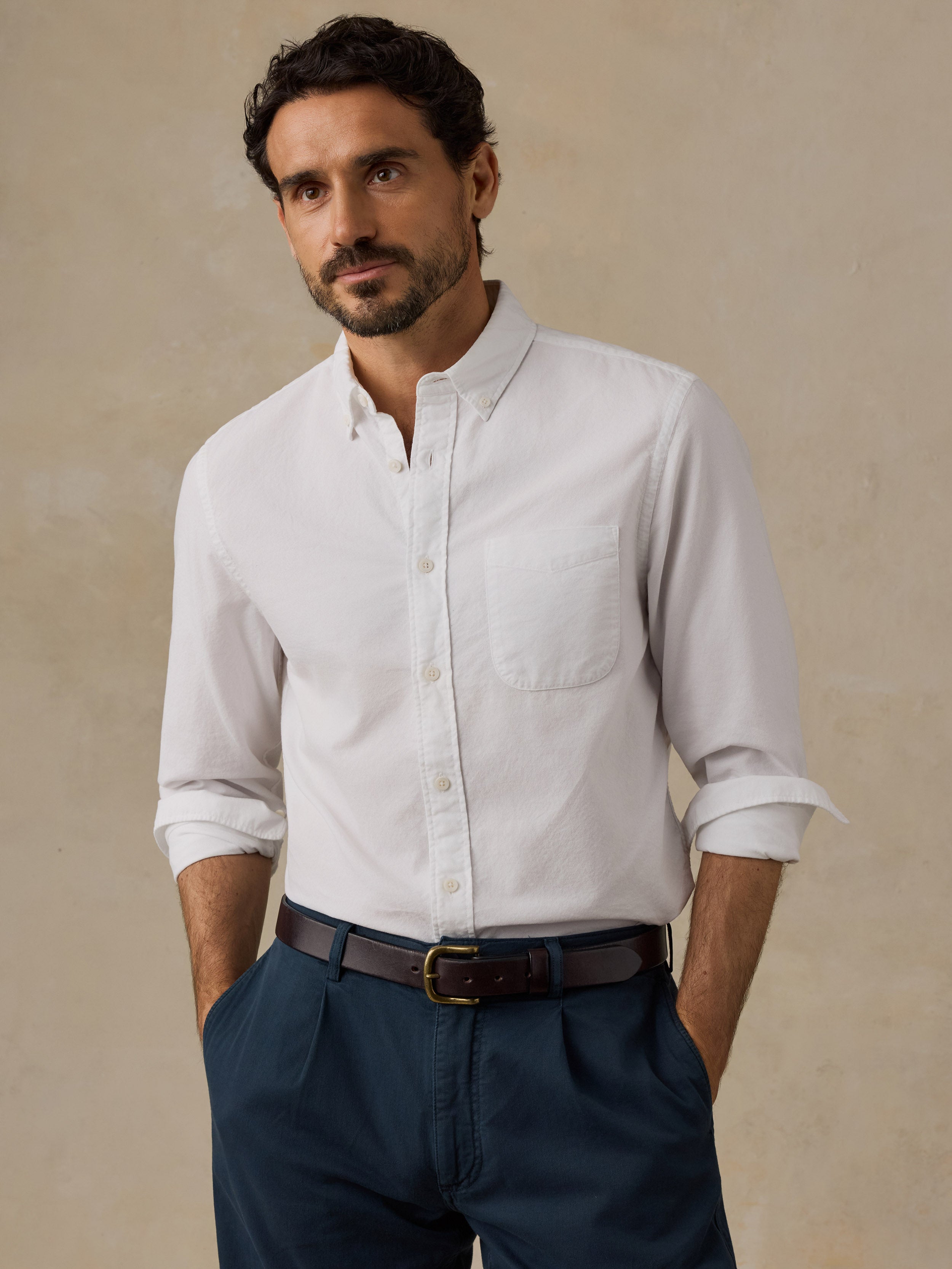 A man wearing a white long-sleeve button-down shirt with rolled-up sleeves, paired with navy blue trousers and a dark brown belt, standing against a textured beige background.