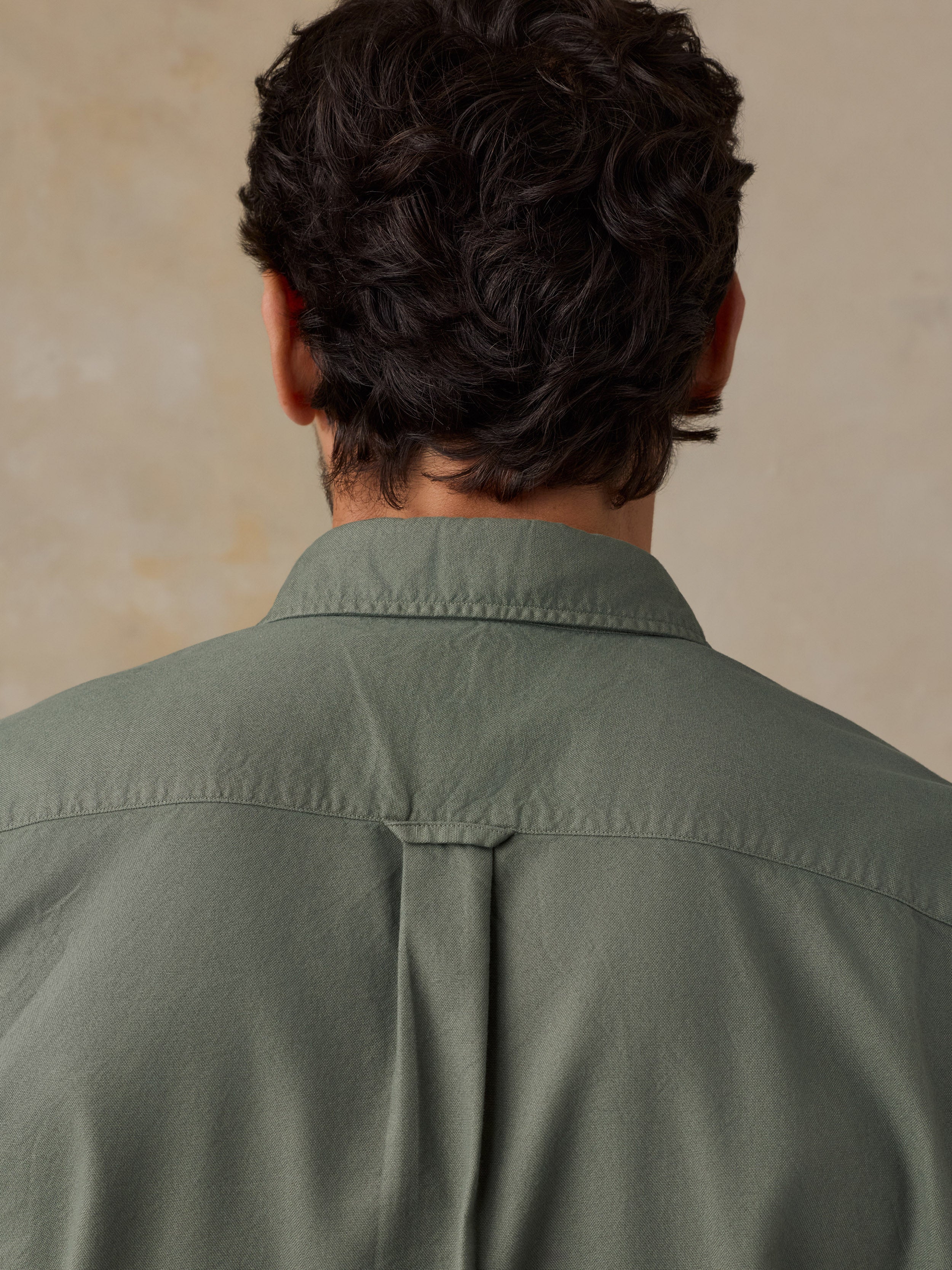 Back view of a man wearing an olive green button-down shirt, showing the collar, shoulder seams, and a central back pleat, set against a textured beige background.