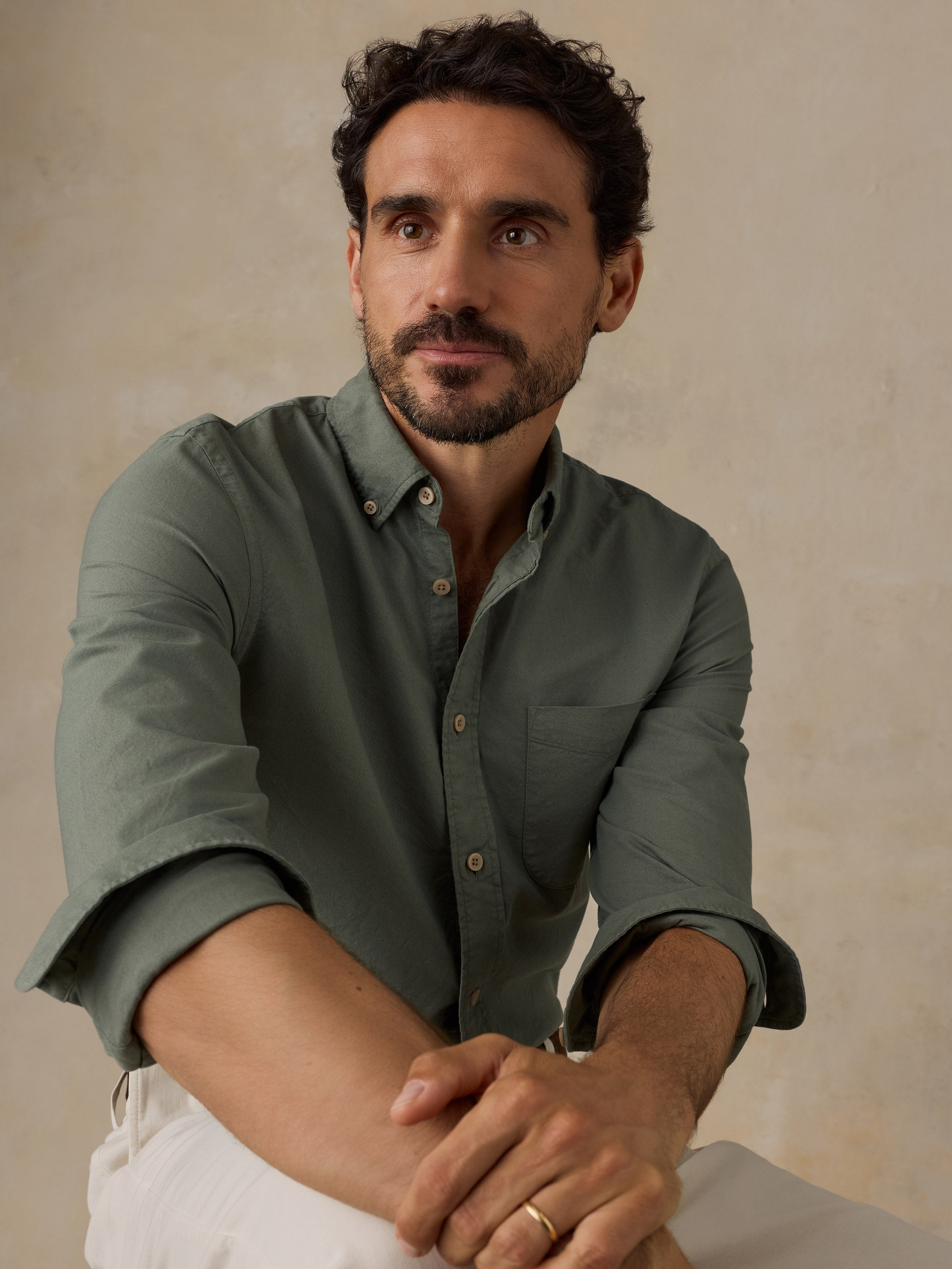 A man wearing a muted sage green long-sleeve button-down shirt with rolled-up sleeves and white trousers, posed against a neutral textured beige background.