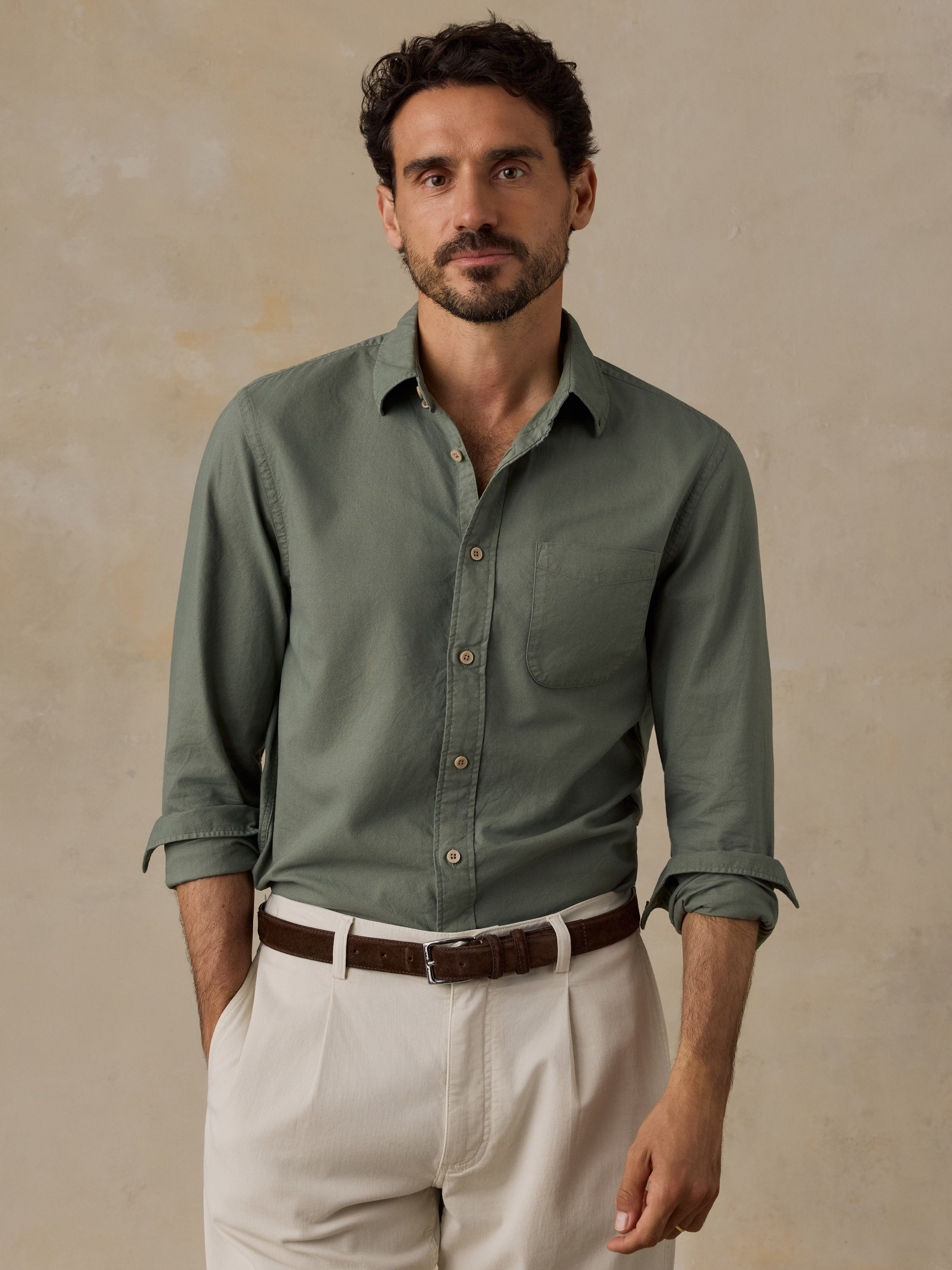 A man wearing a sage green long-sleeve button-down shirt with rolled-up sleeves, paired with off-white trousers and a brown belt, standing against a textured beige background.