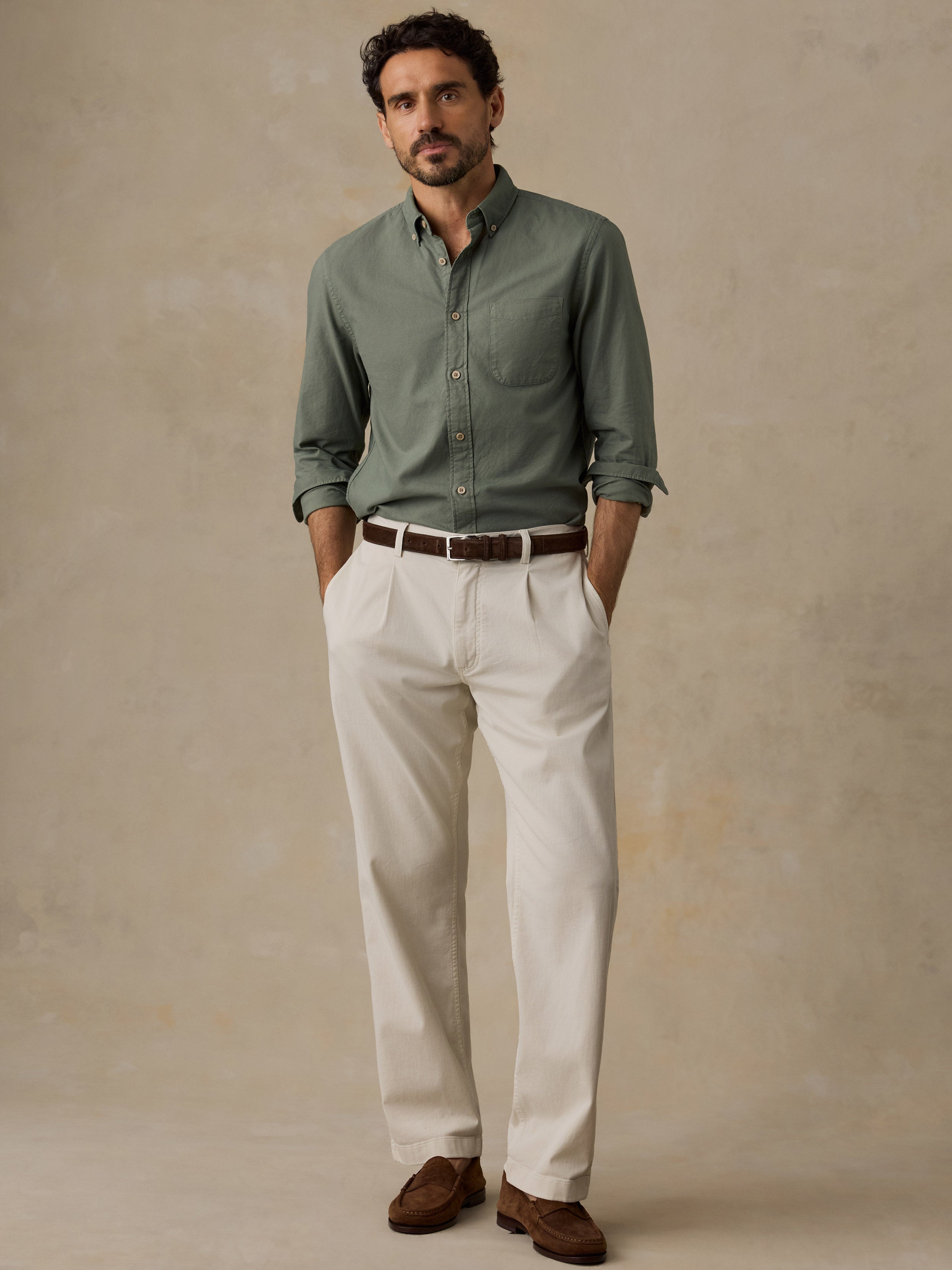 A man wearing a sage green button-down shirt, off-white trousers, a brown belt, and brown leather shoes, standing against a textured beige studio backdrop.