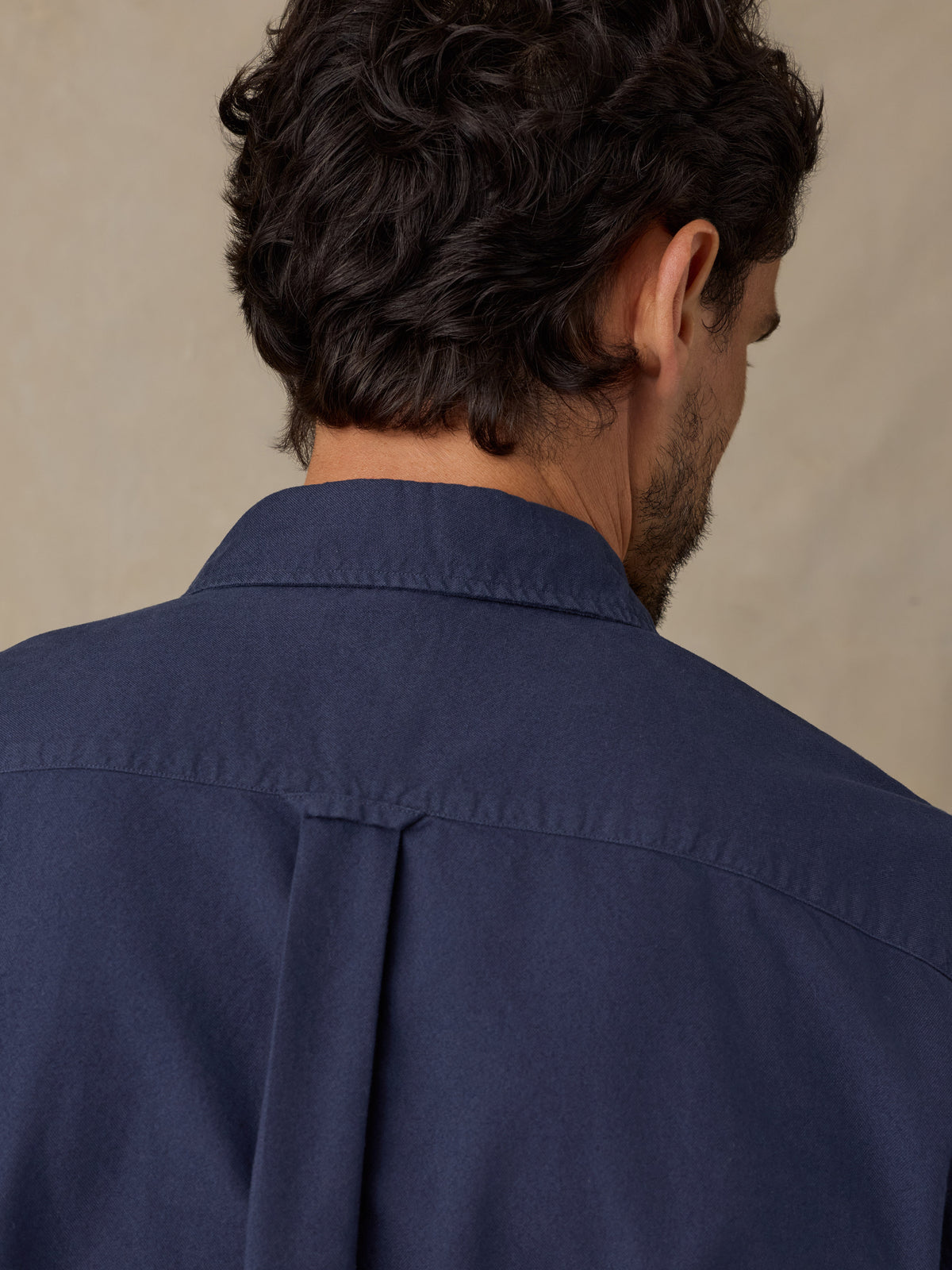 Back view of a man wearing a navy blue button-down shirt, showing the collar, shoulder, and back fabric against a neutral beige background., image 4 of 9