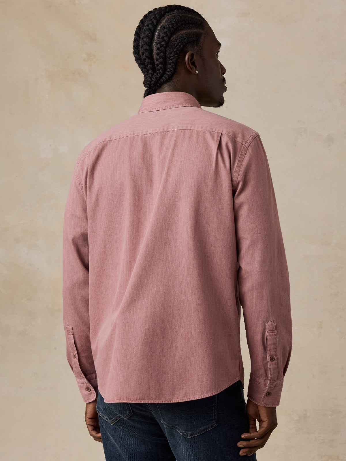 A man viewed from behind wearing a dusty rose pink long-sleeve button-down shirt and dark blue jeans, standing against a textured beige wall., image 3 of 6