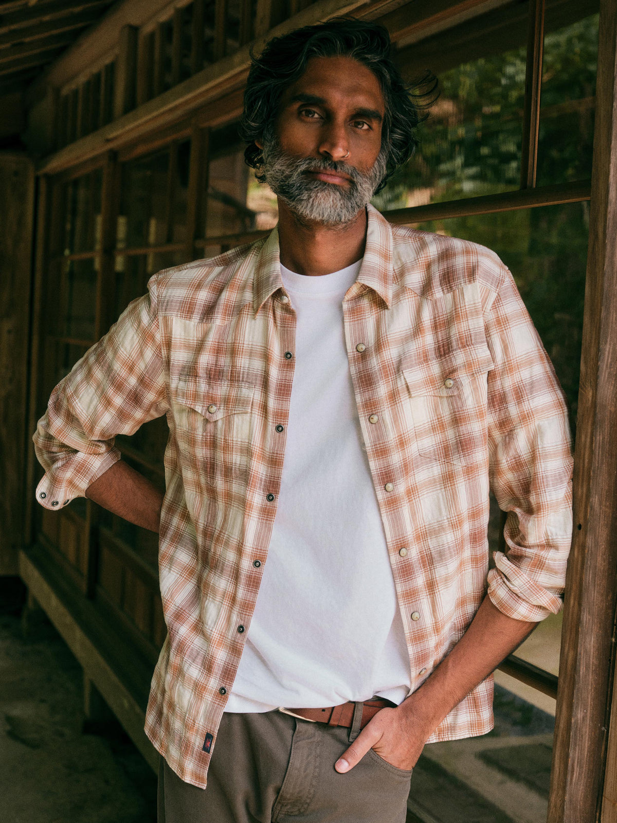 A man with a graying beard and dark hair stands on a rustic wooden porch wearing a light beige and brown plaid button-down shirt over a white t-shirt and khaki pants., image 1 of 8