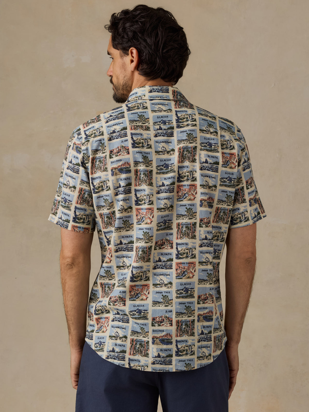 A man viewed from the back wearing a short-sleeve button-up shirt with an all-over collage print of vintage harbor scenes, paired with navy pants, against a textured beige studio background., image 3 of 7