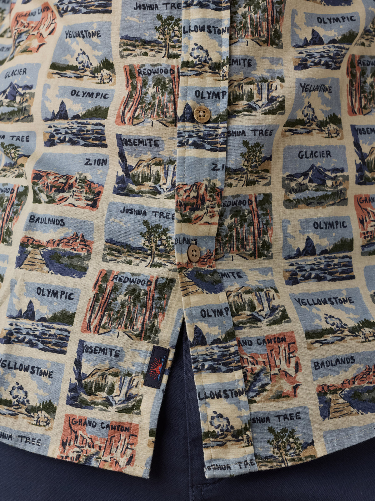 A close-up of a button-front garment, likely a shirt or jacket, featuring an all-over printed pattern of illustrated canyon and mountain scenes with labels such as Yellowstone, Grand Canyon, Zion, and Olympic. The print is set on a light cream background with blue, red, green, and brown scenic illustrations., image 4 of 7