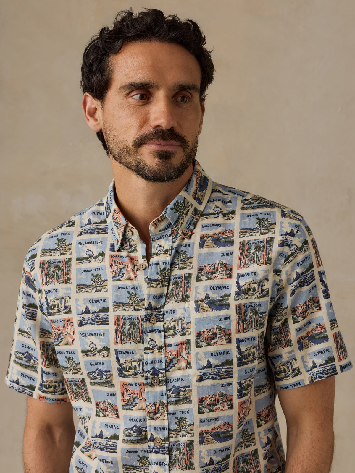 A man wearing a short-sleeve button-up shirt with a vintage postcard-style collage print featuring coastal scenes and text, posed against a neutral beige background., image 2 of 7