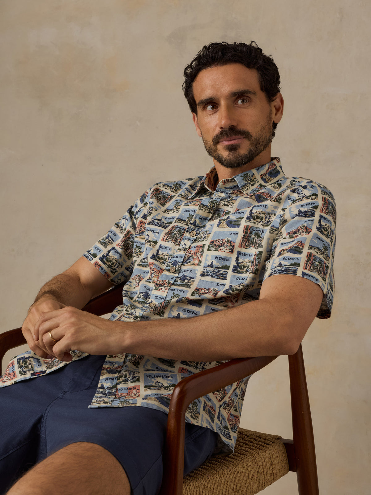 A man wearing a short-sleeve button-up shirt with a detailed scenic print, seated in a wooden chair against a textured beige wall., image 6 of 7