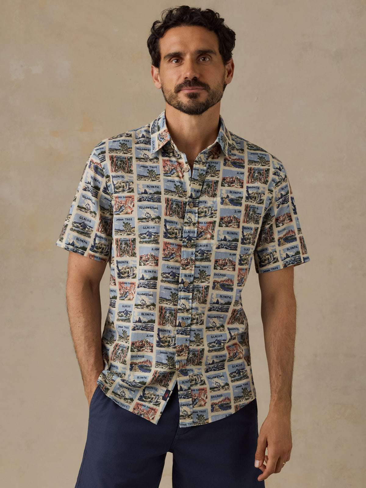 A man wearing a short-sleeve button-up shirt with a detailed scenic print, paired with navy blue pants, standing against a textured beige background., image 1 of 7