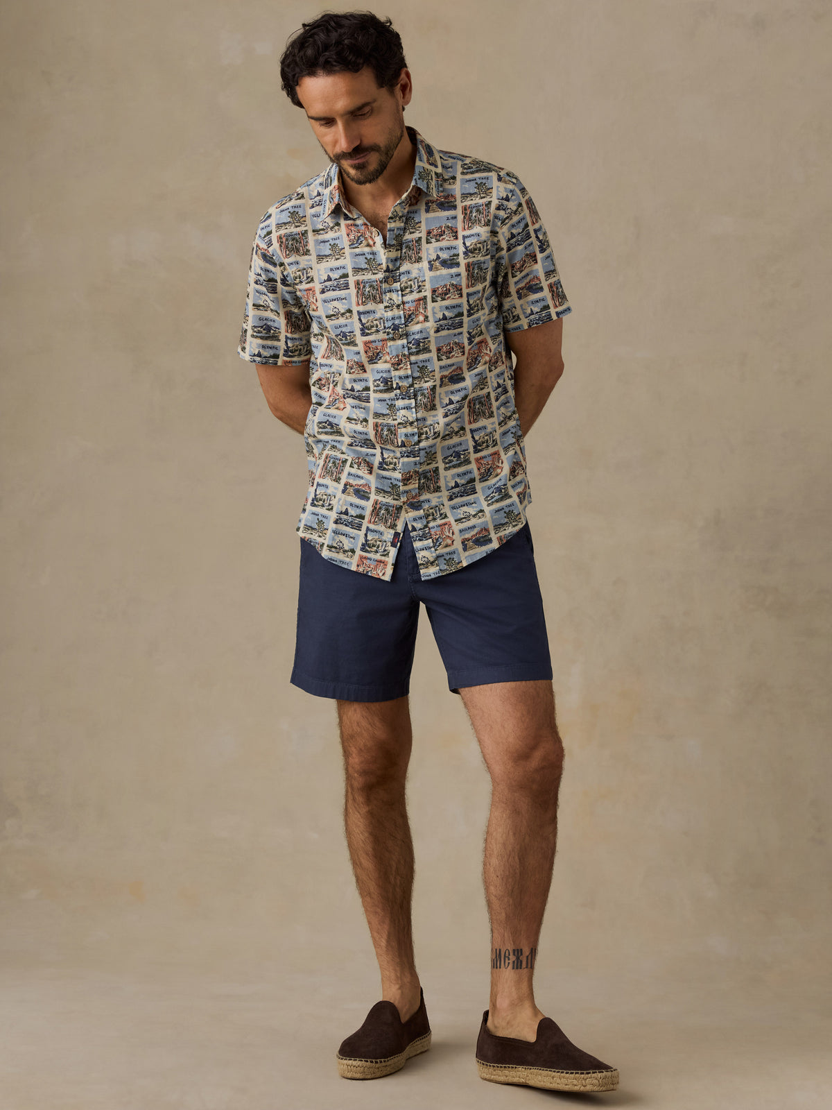 A man wearing a short-sleeve button-up shirt with a detailed scenic print, navy blue shorts, and brown espadrille loafers, standing against a textured beige studio backdrop., image 5 of 7