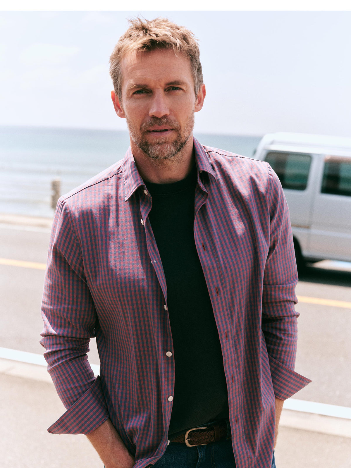 A man standing outdoors near the ocean wearing a purple and red striped button-down shirt over a black t-shirt, with a white van in the background., image 7 of 9