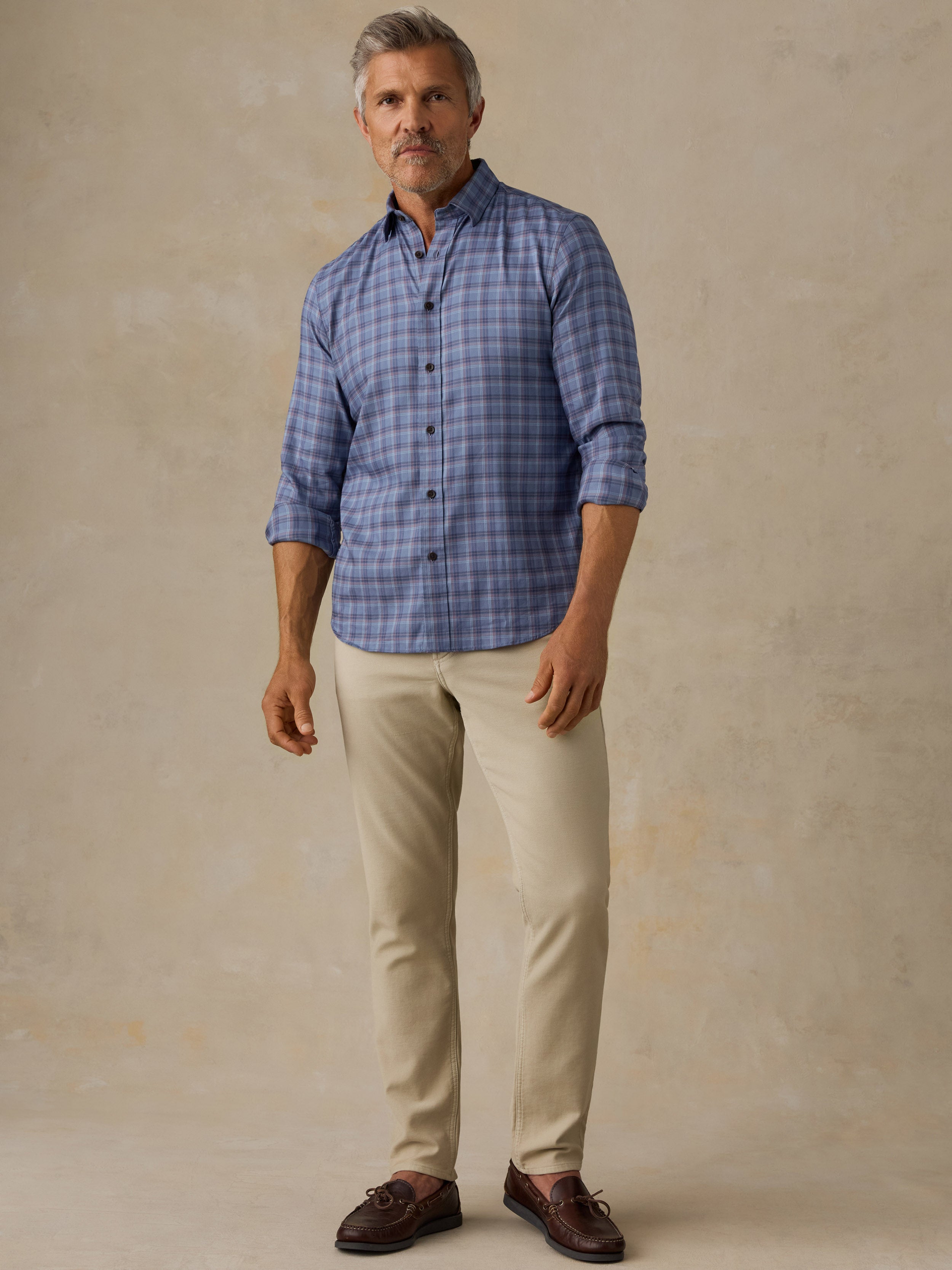 A man modeling a blue grey plaid button-down shirt with beige trousers and brown leather shoes, standing against a textured beige studio backdrop.