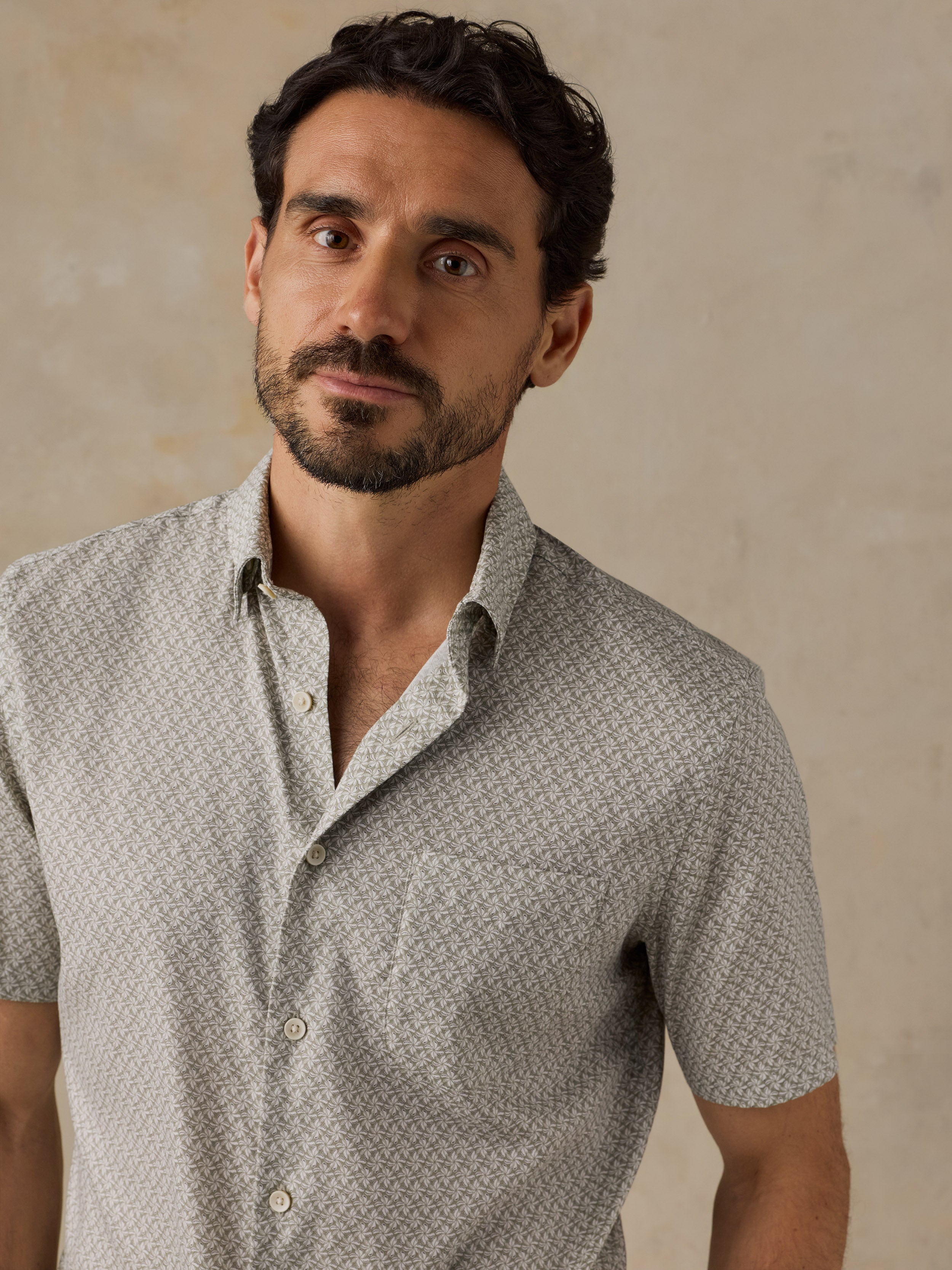 A man wearing a light green short-sleeve button-up shirt with a subtle geometric pattern, photographed against a textured beige background.