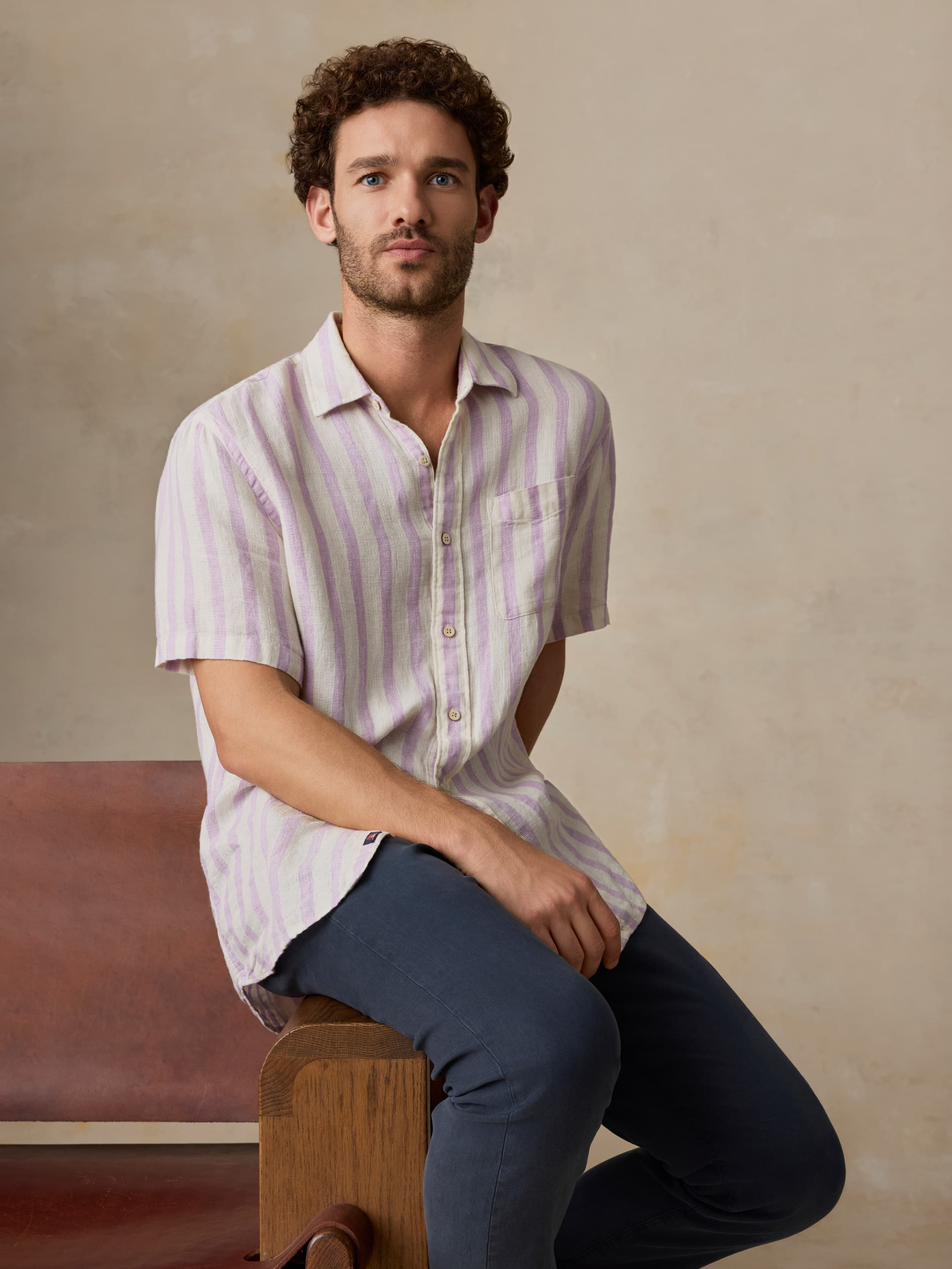 Man sitting on a wooden stool wearing a striped shirt and dark pants against a beige background
