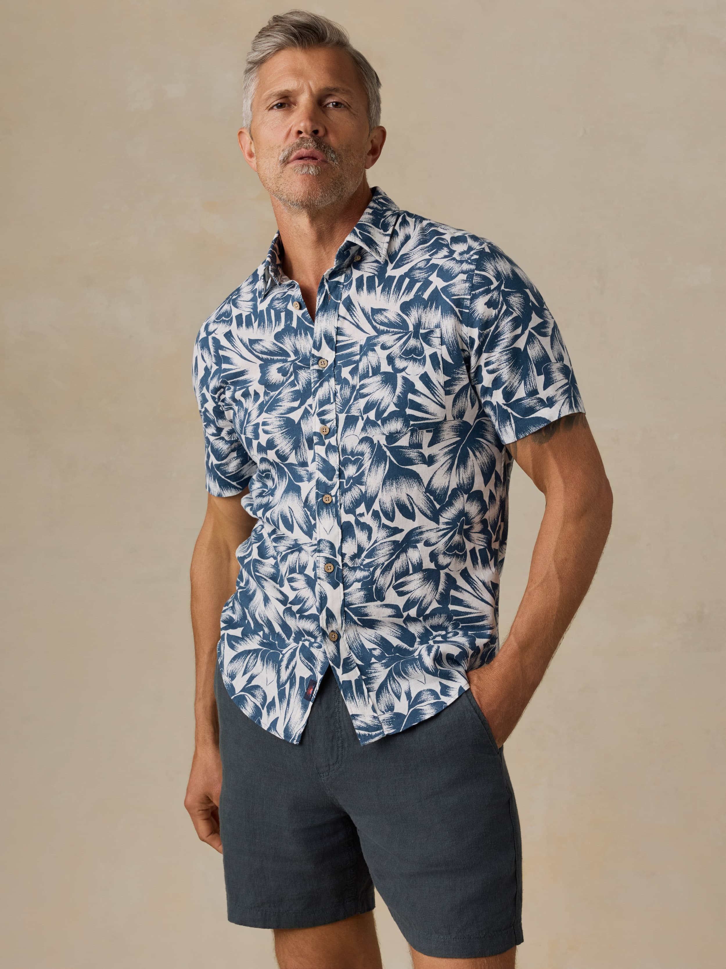 Man wearing a blue and white patterned shirt with navy shorts against a beige background