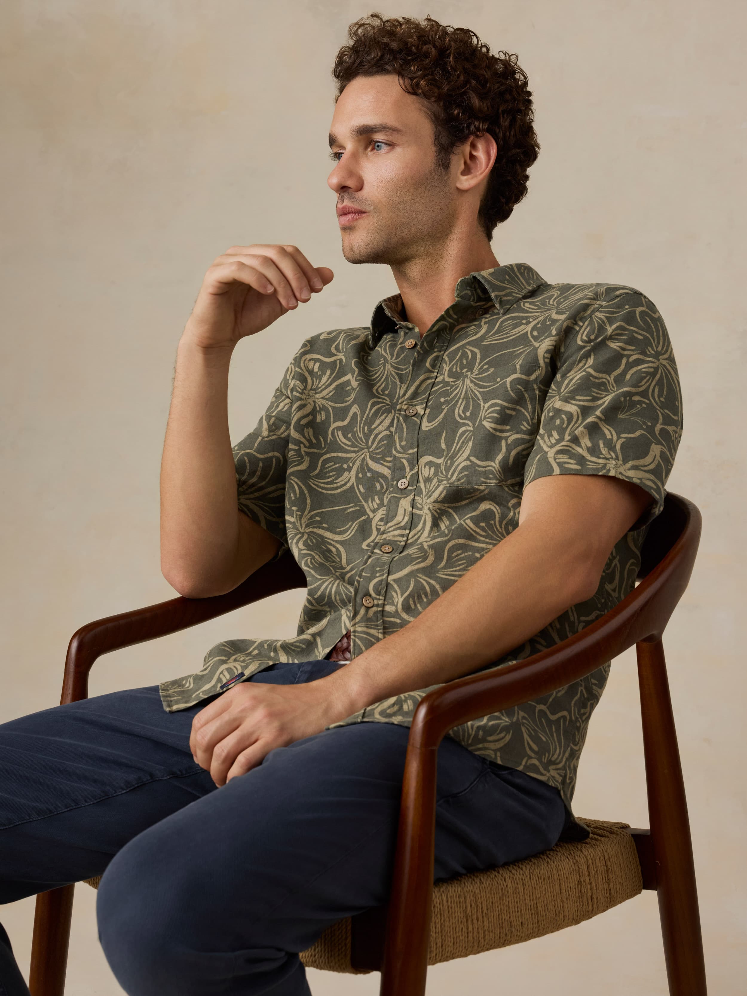 Man sitting on a wooden chair wearing a green floral printed shirt and navy pants against a beige background