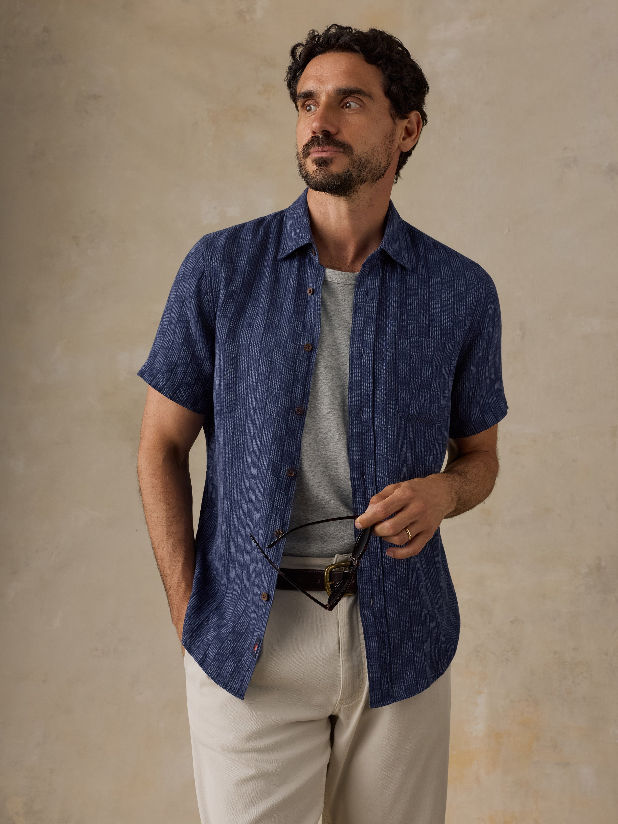 A man wearing a short-sleeve navy blue textured button-up shirt over a gray t-shirt and beige trousers, holding a pair of glasses against a textured beige background.