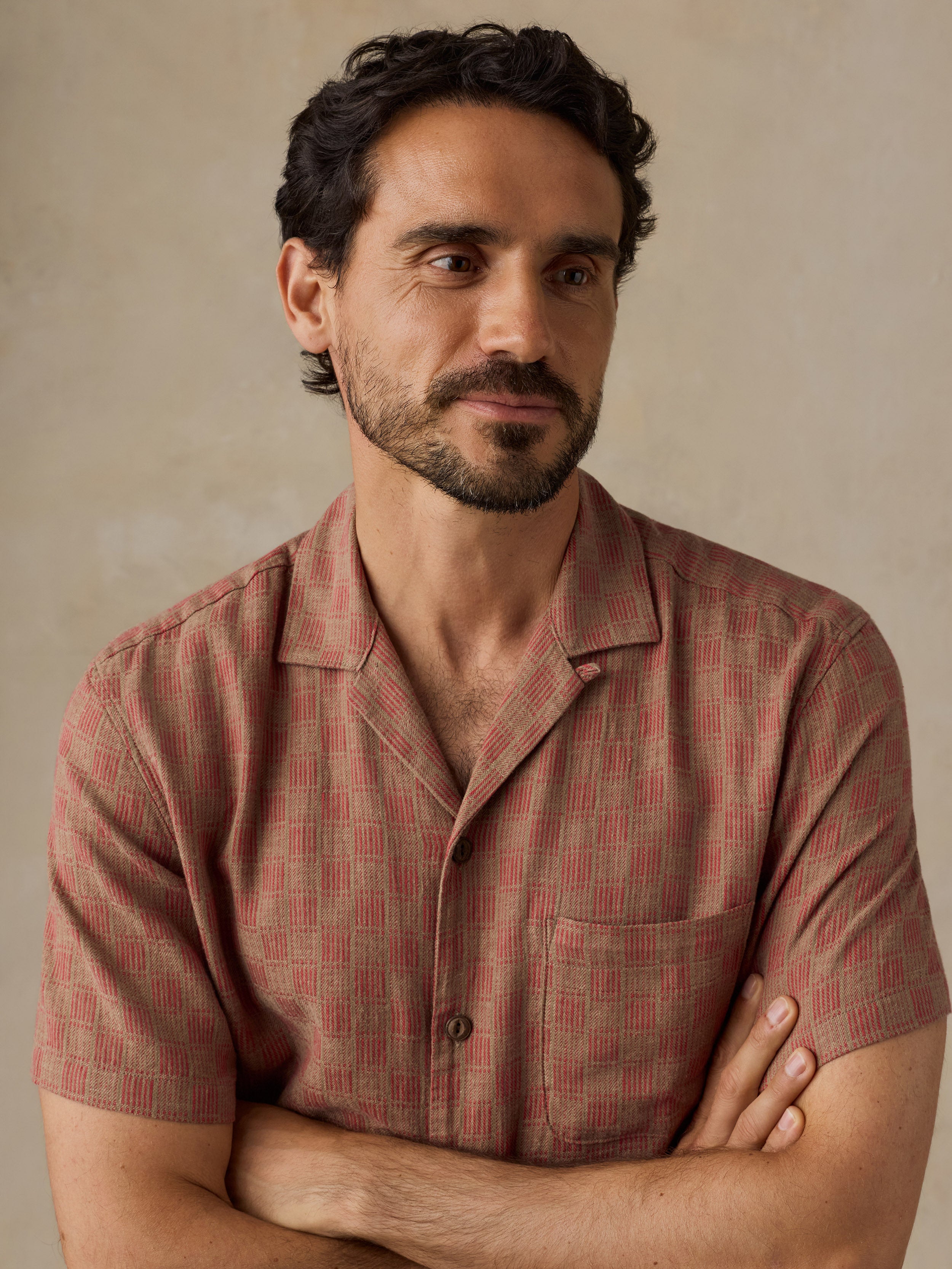 A man with dark curly hair and a beard wearing a short-sleeve button-up shirt with a red and beige geometric pattern, posed with arms crossed against a neutral beige background.