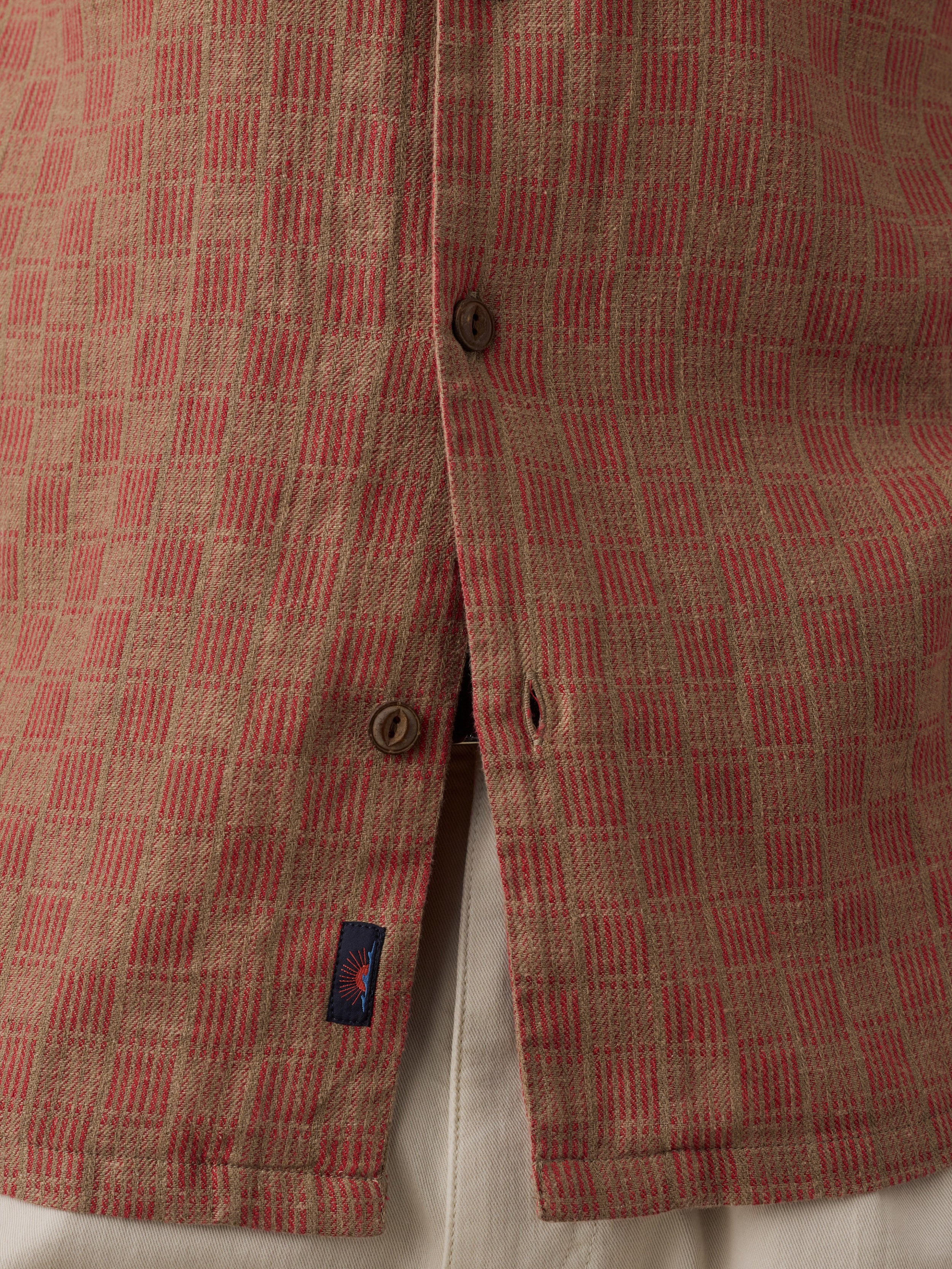 Close-up of a textured woven shirt fabric with a red and beige basketweave pattern, featuring brown buttons and a small Faherty brand tag.