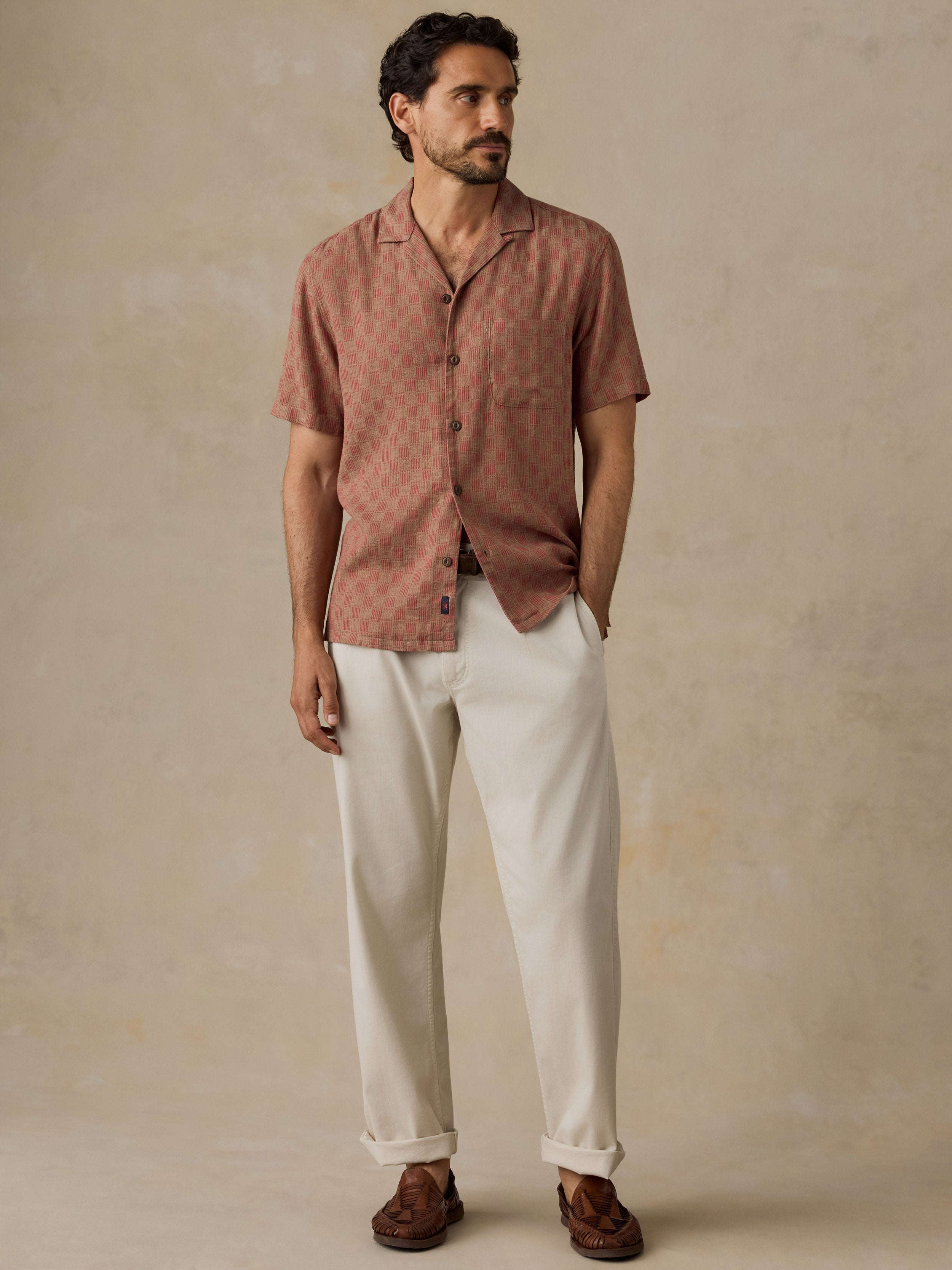 A man wearing a short-sleeve button-up shirt in a dusty rose-brown color with a subtle geometric pattern, paired with off-white trousers and brown shoes, standing against a textured beige background.
