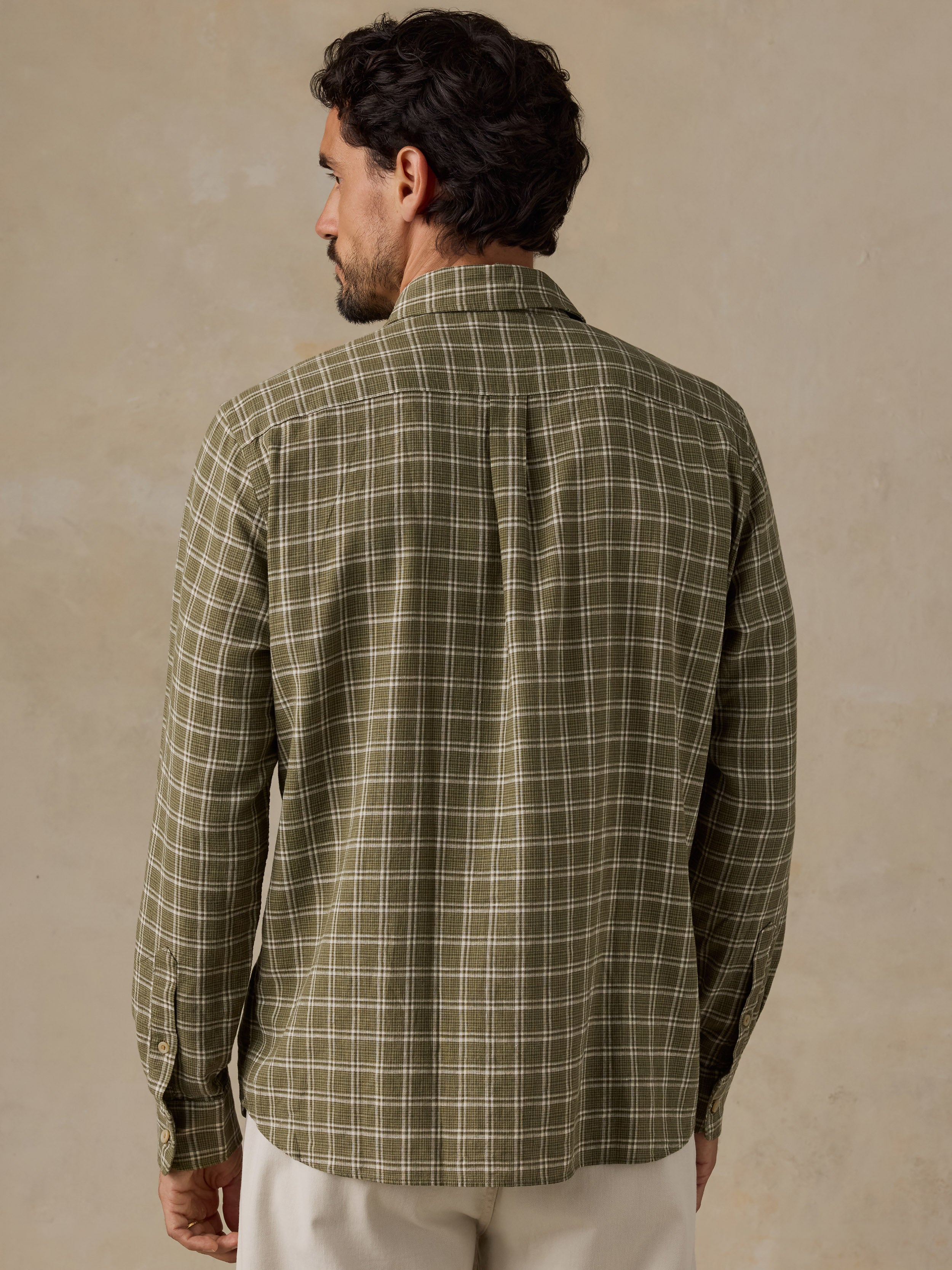 A man shown from the back wearing a long-sleeve plaid button-up shirt in olive green and beige, paired with off-white trousers, against a textured beige background.
