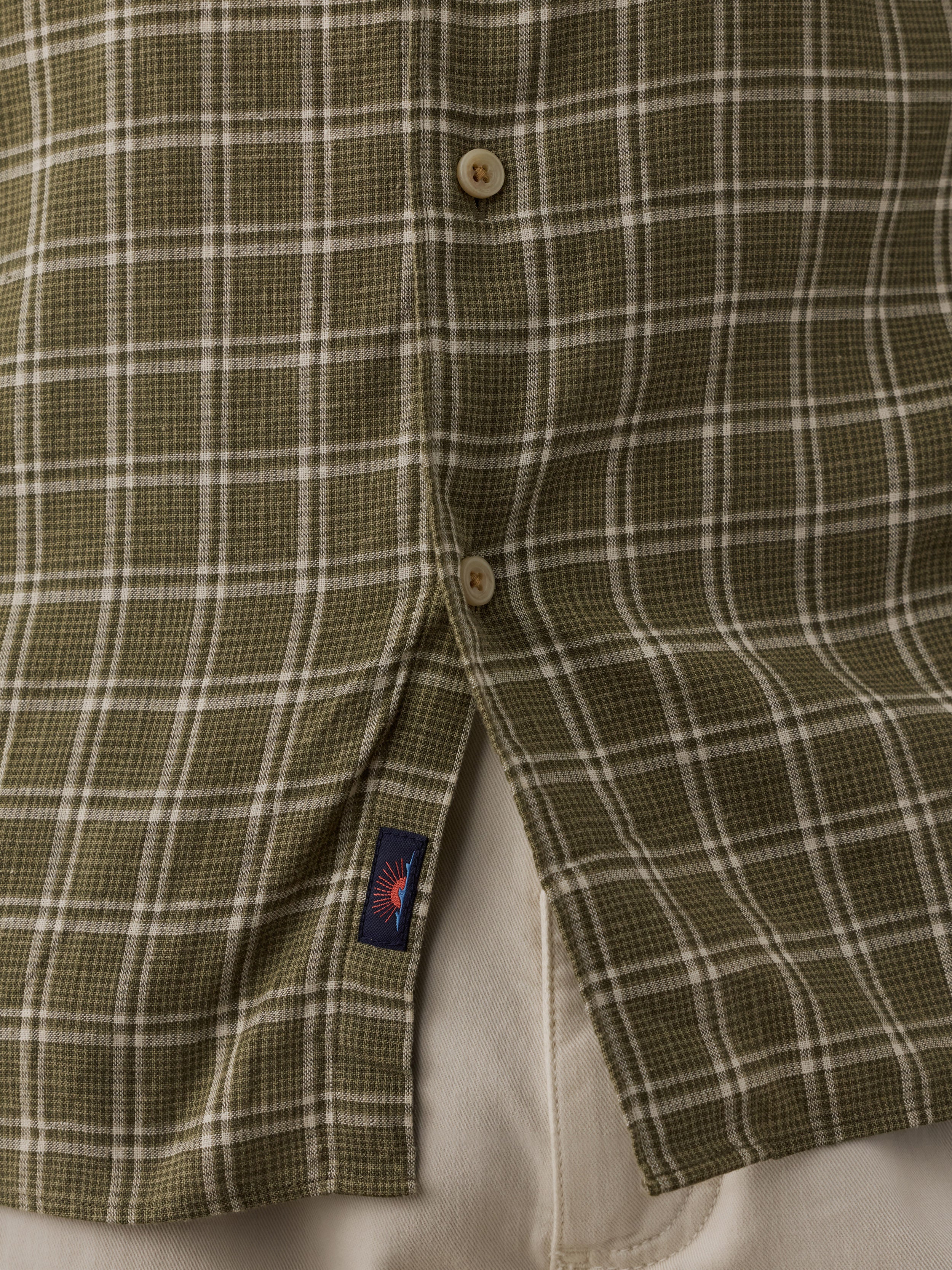 Close-up of a plaid button-up shirt in olive green and white, showing the front buttons and a small Faherty brand tag near the hem.