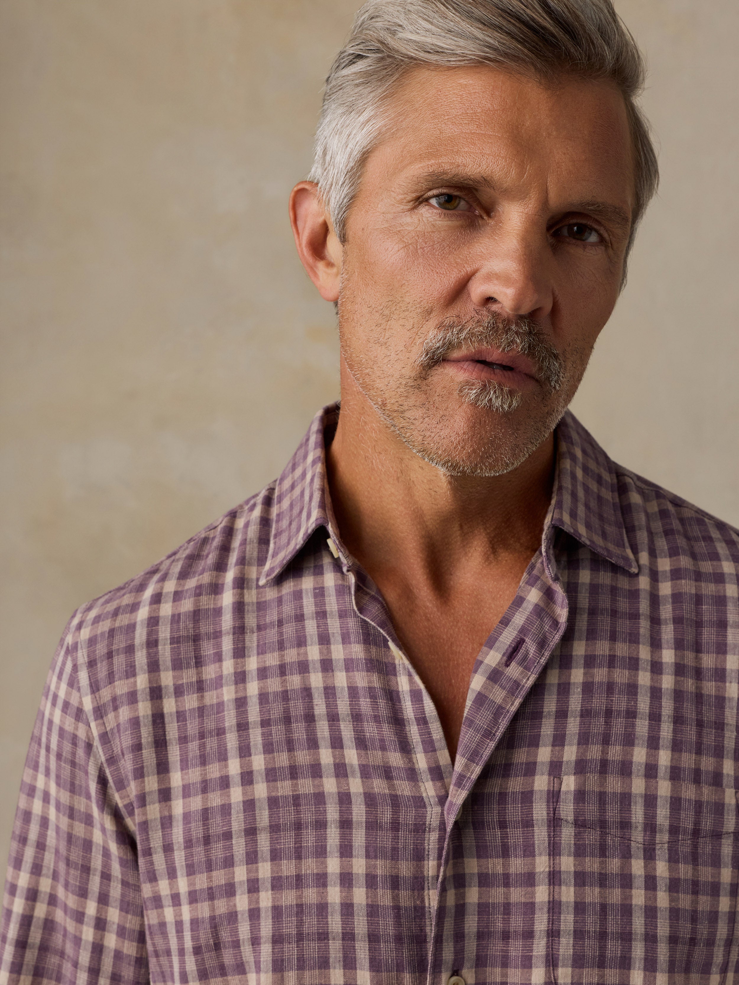 A close-up portrait of a man with gray hair and a mustache wearing a purple and beige plaid button-down shirt against a textured beige background.