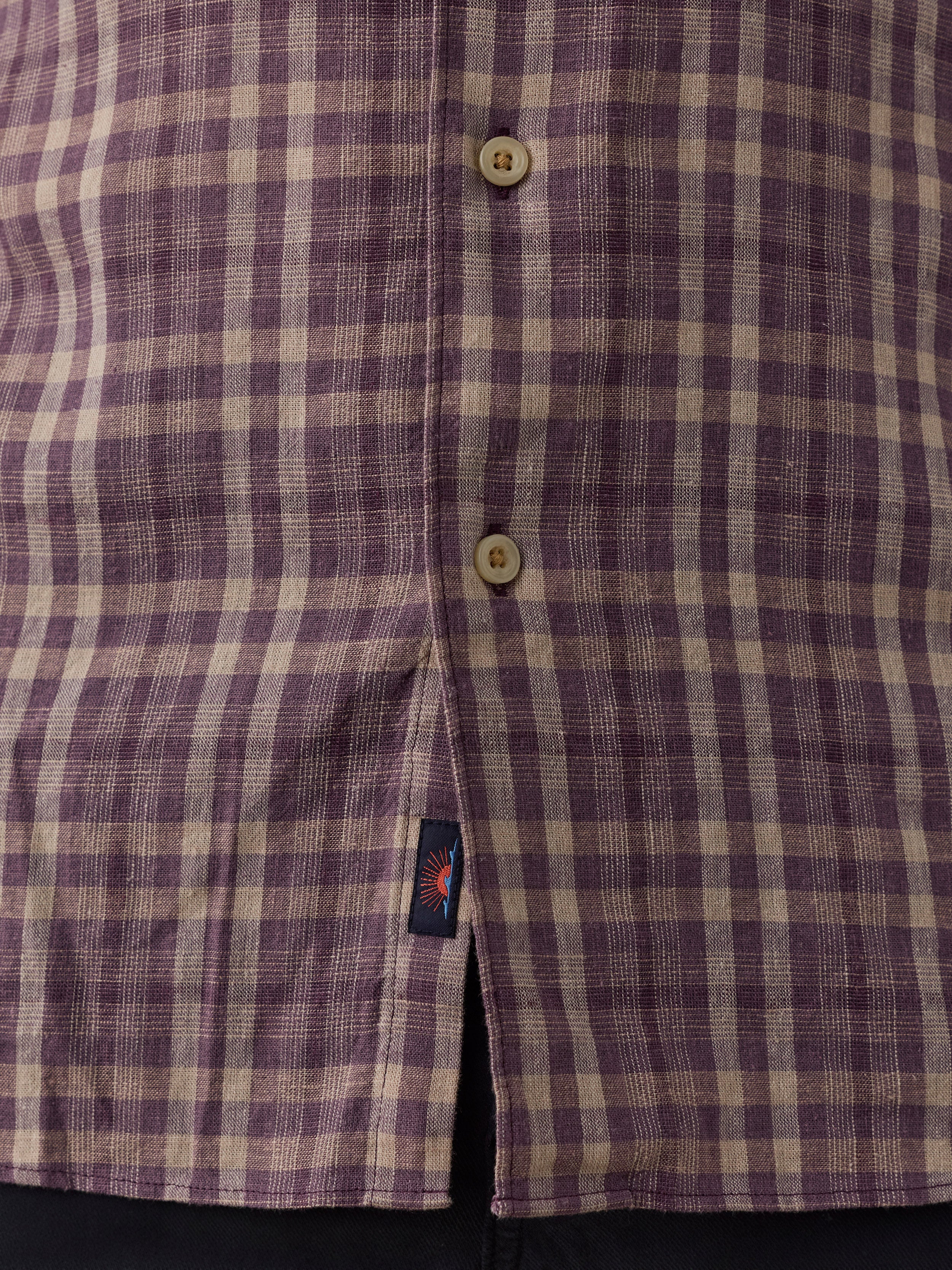 Close-up of a purple and beige plaid button-down shirt fabric, showing the button placket and a small Faherty brand tag near the hem.