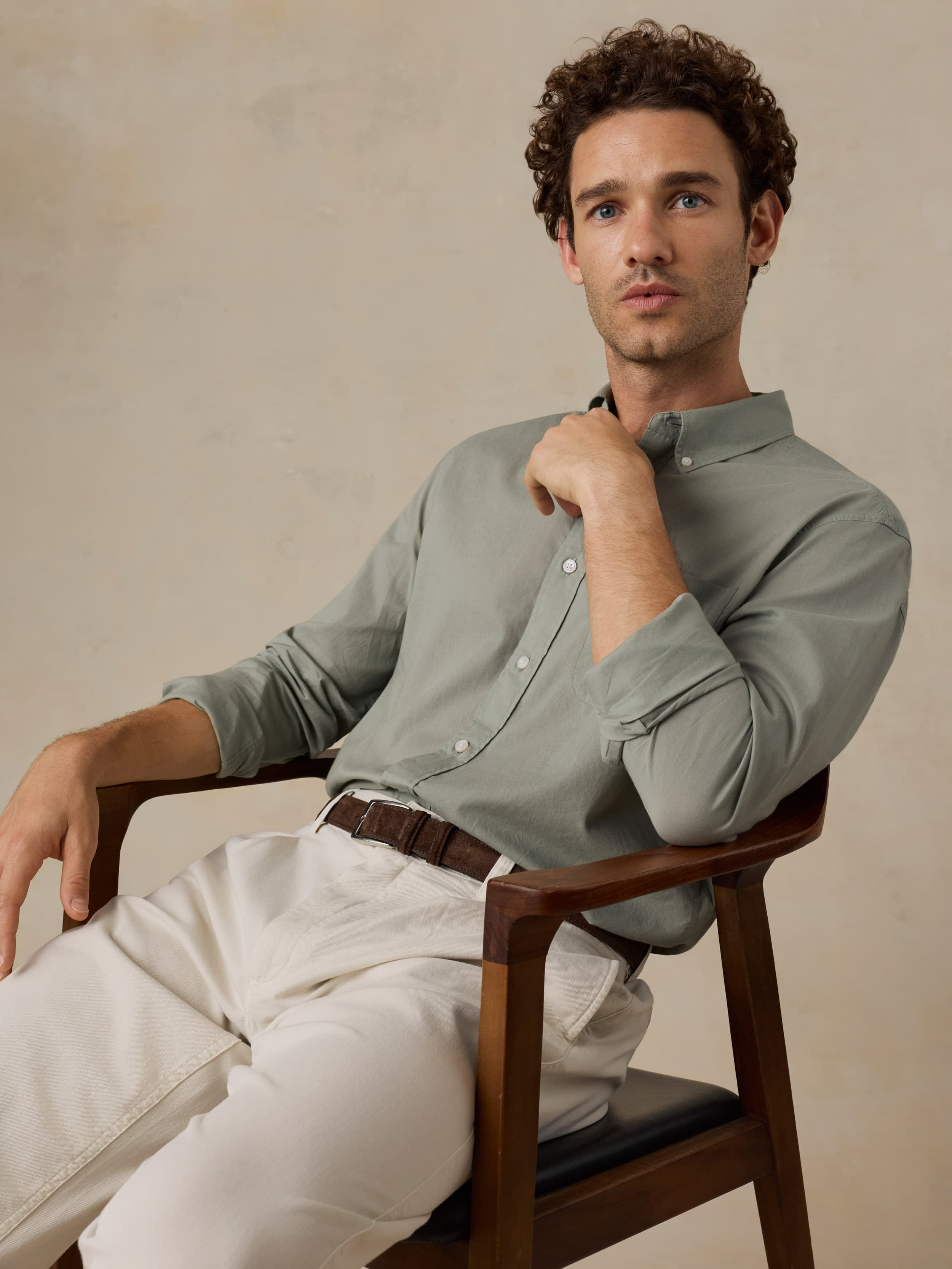 Man sitting on a wooden chair wearing a light green shirt and white pants against a beige background