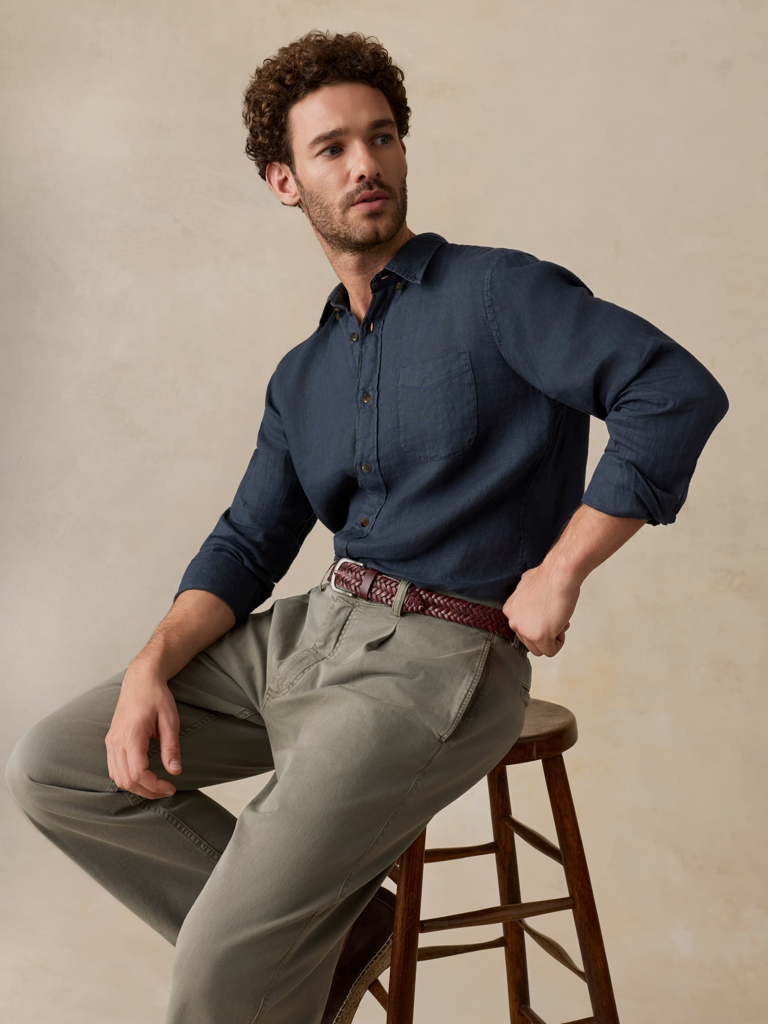 Man sitting on a wooden stool wearing a blue shirt and beige pants against a beige background
