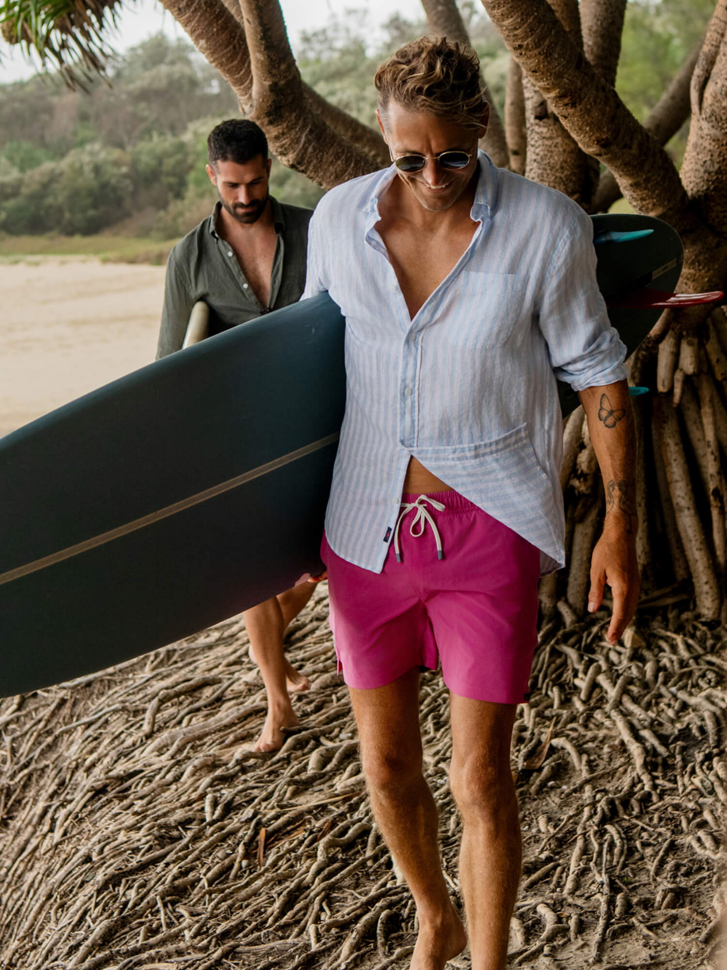 Man in pink swim trunks and a striped linen shirt holding a surfboard on a beach