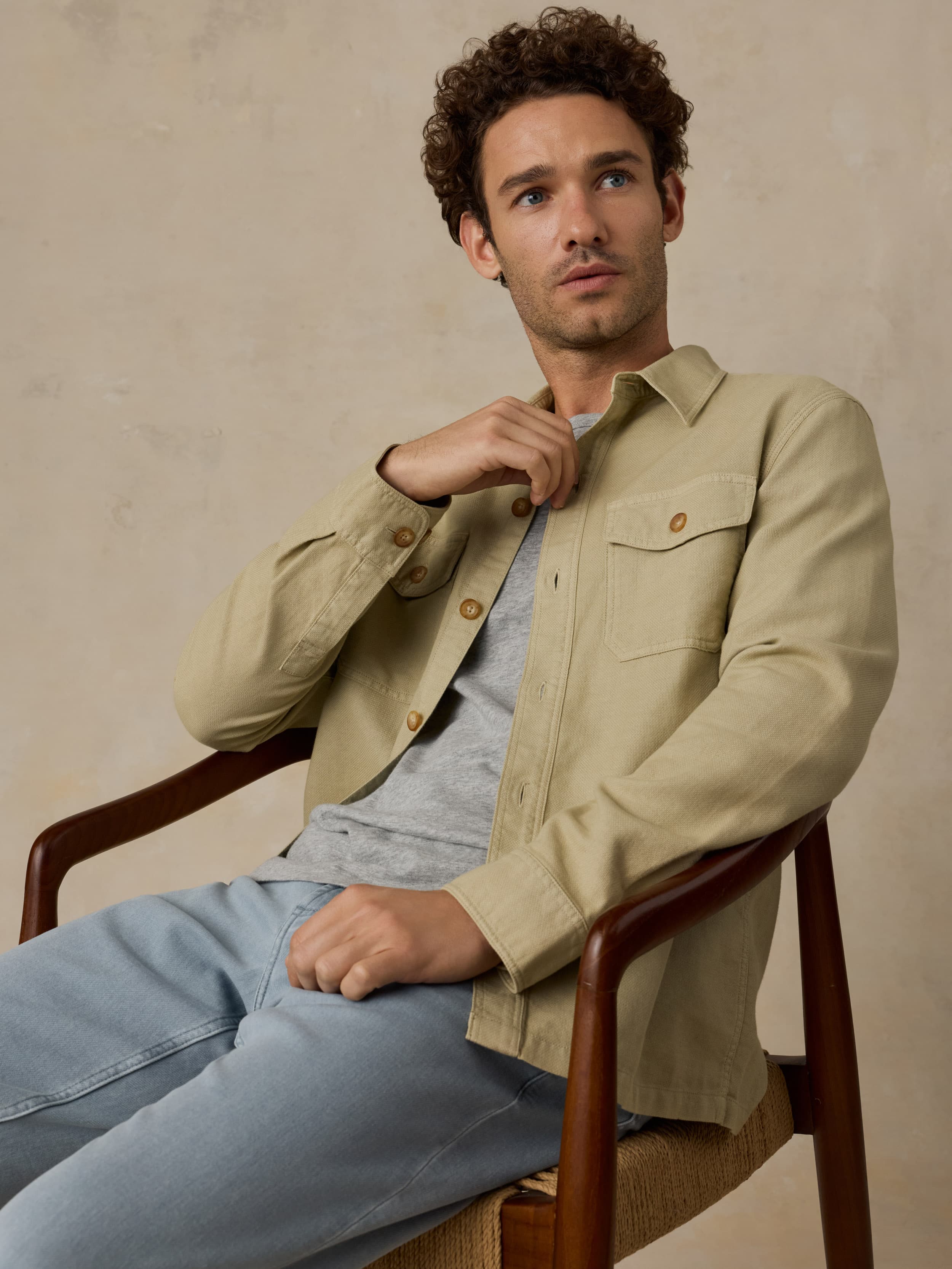 Man sitting on a wooden chair wearing a beige button down shirt with blue jeans against a beige background