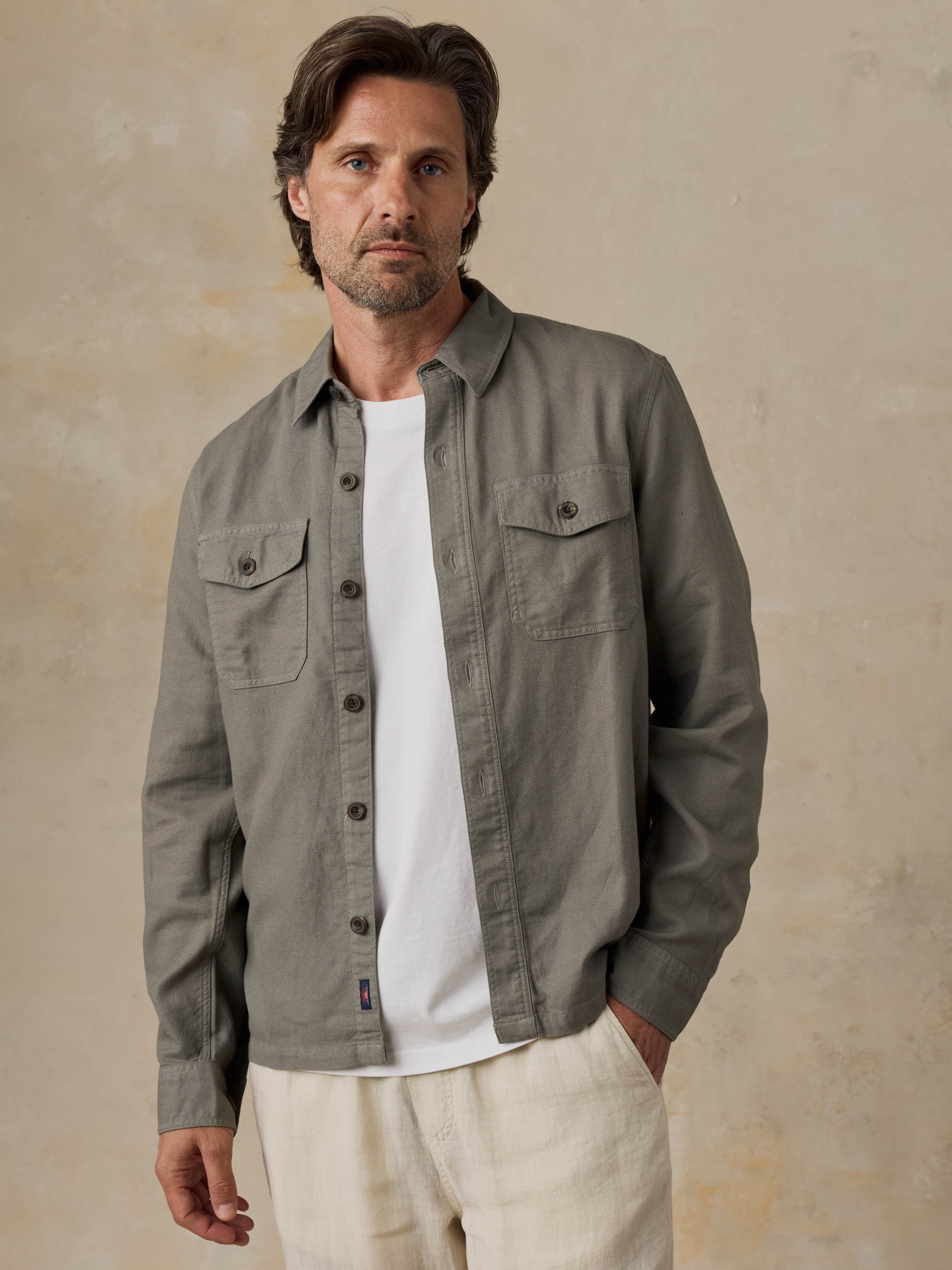Man wearing an olive green jacket over a white shirt with beige pants against a beige background