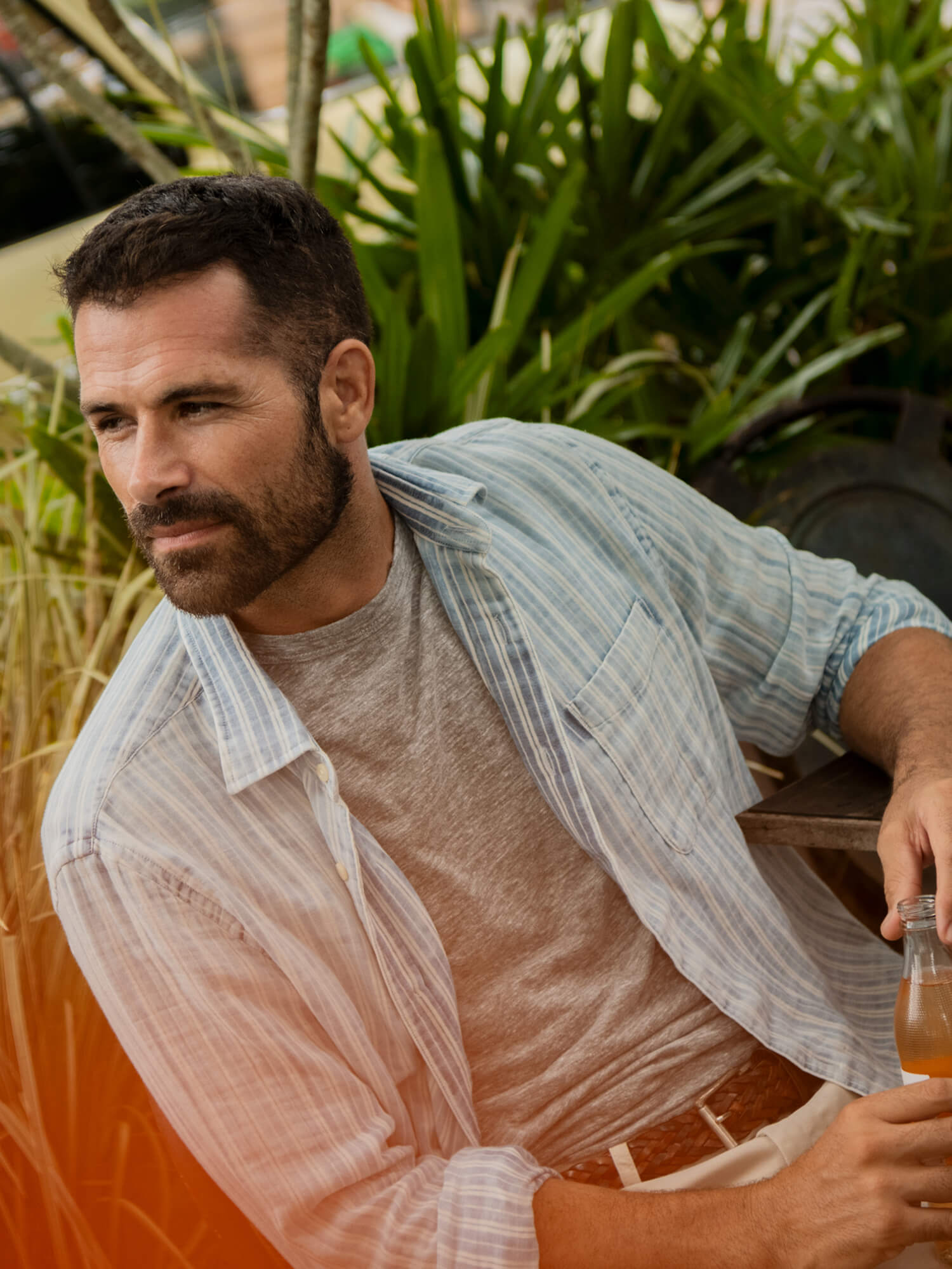 Man wearing a striped shirt layered over a grey t-shirt sitting on a chair outside