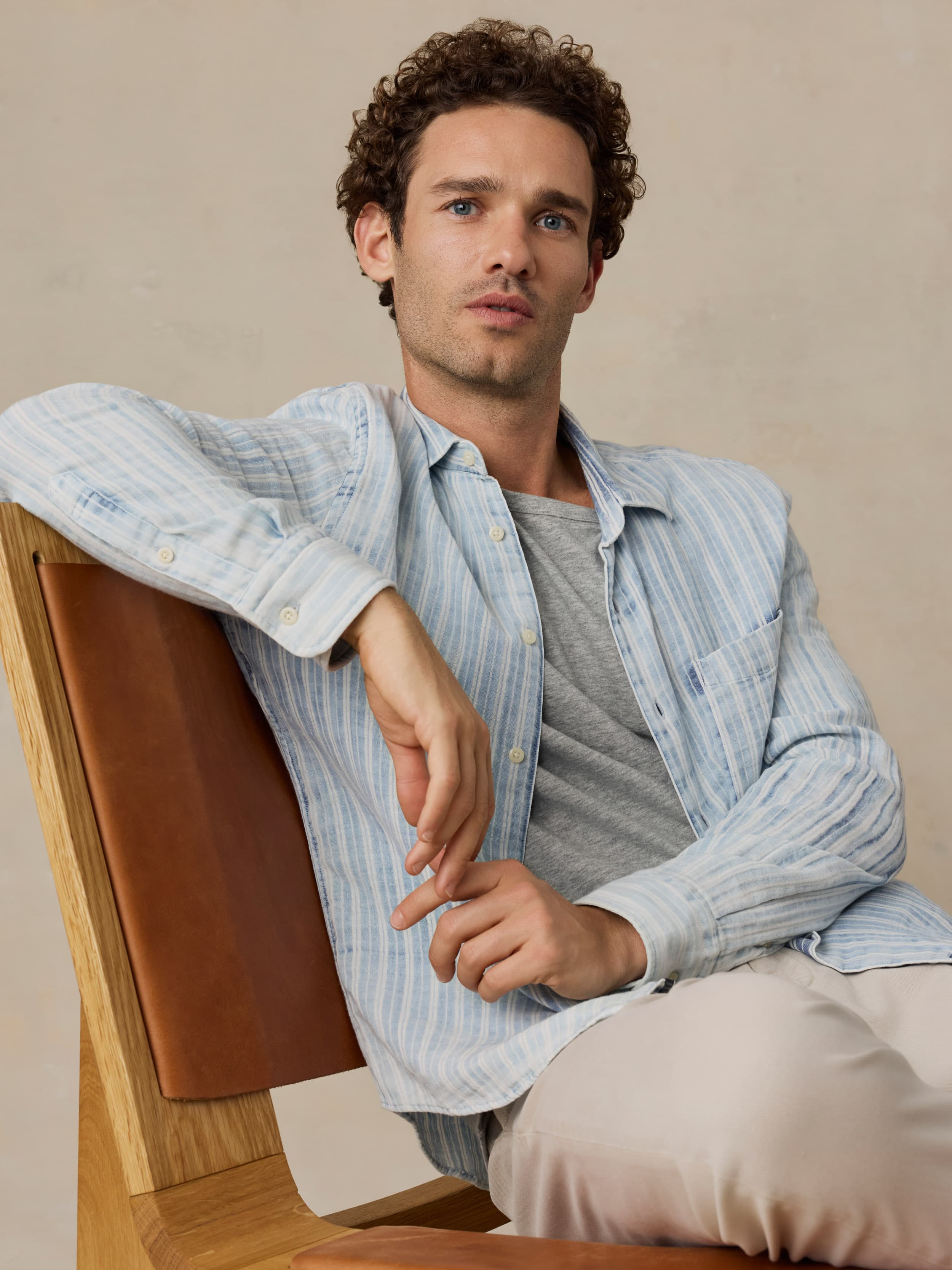 Man sitting on a wooden chair wearing a light blue striped shirt over a gray t-shirt.